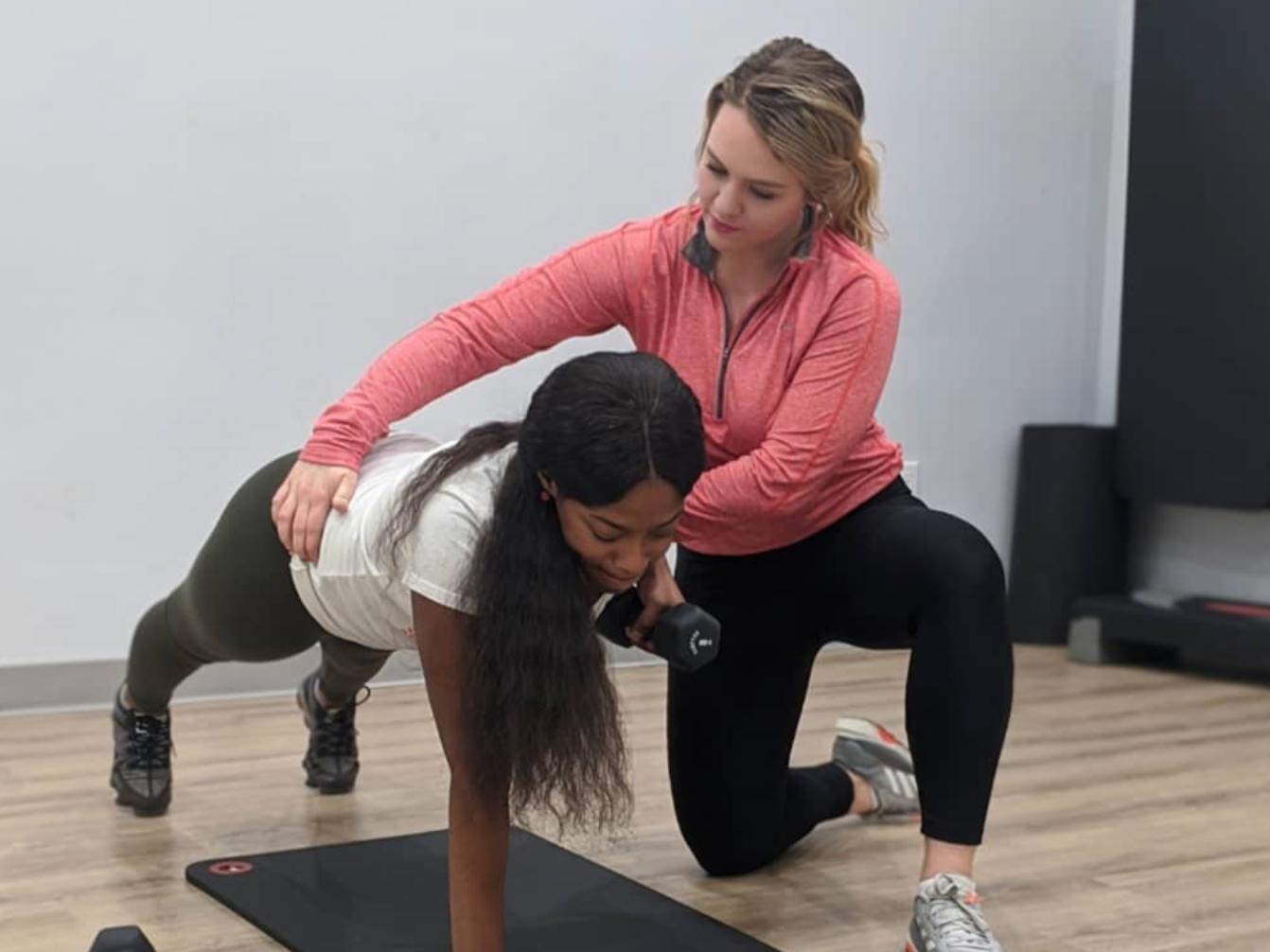 A woman is assisting another woman with her workout in a gym; the woman being assisted is on all fours, holding a dumbbell, while the trainer supports her by placing a hand on her back and her knee.