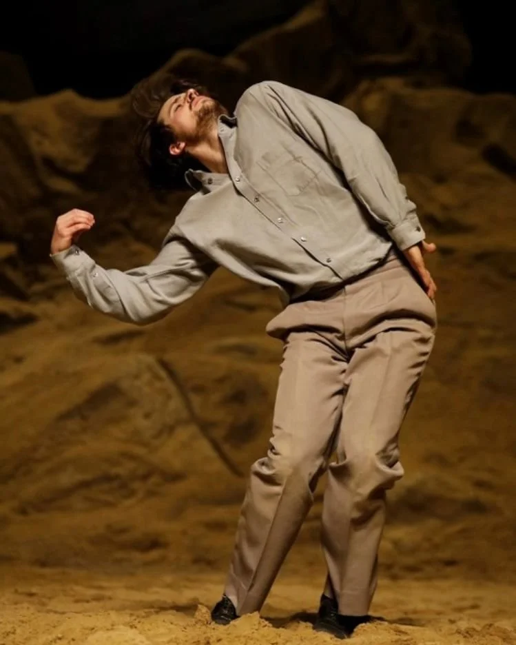 A man with long hair and a beard is striking a playful pose in a desert-like setting with rocky formations in the background, wearing a beige shirt and pants.