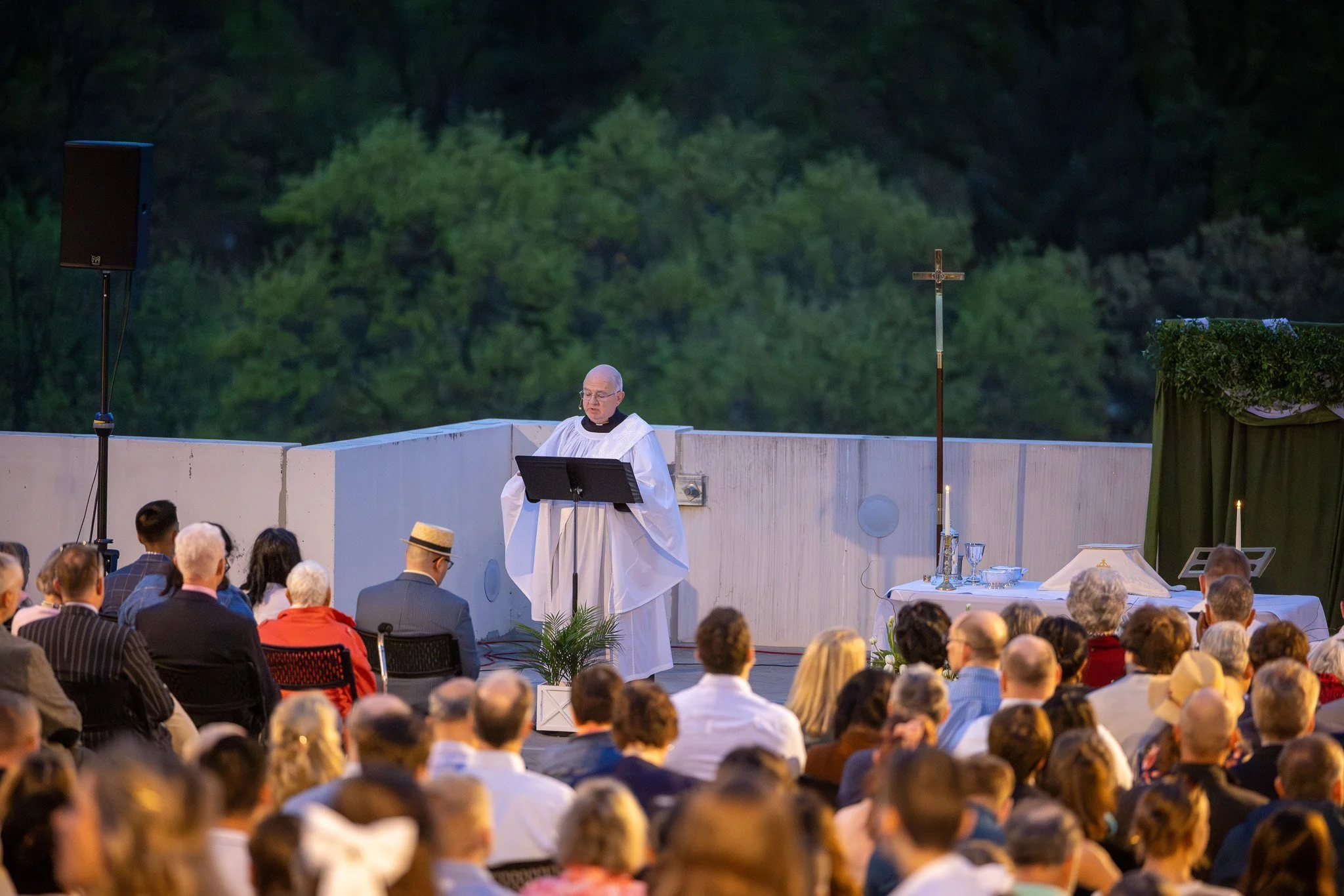 A priest dressed in white robes conducting a religious service outdoors with a congregation seated in front. The altar has a cross, chalice, and other liturgical items. The background shows green trees.