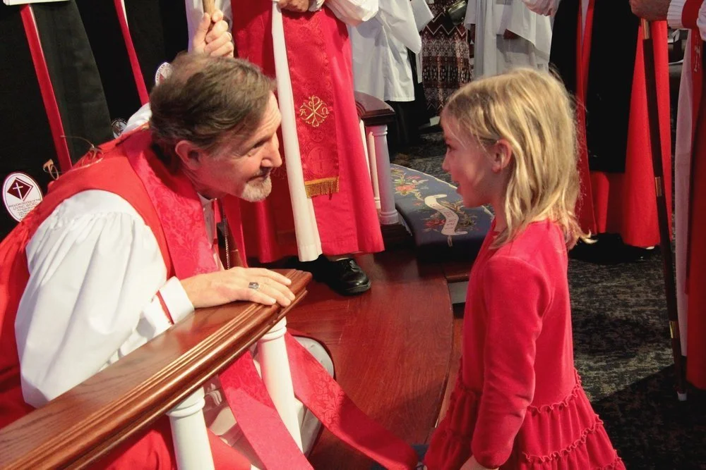 A young girl in a red dress smiling and facing a priest, who is kneeling and leaning over a wooden railing, in a church or religious setting during a ceremony.