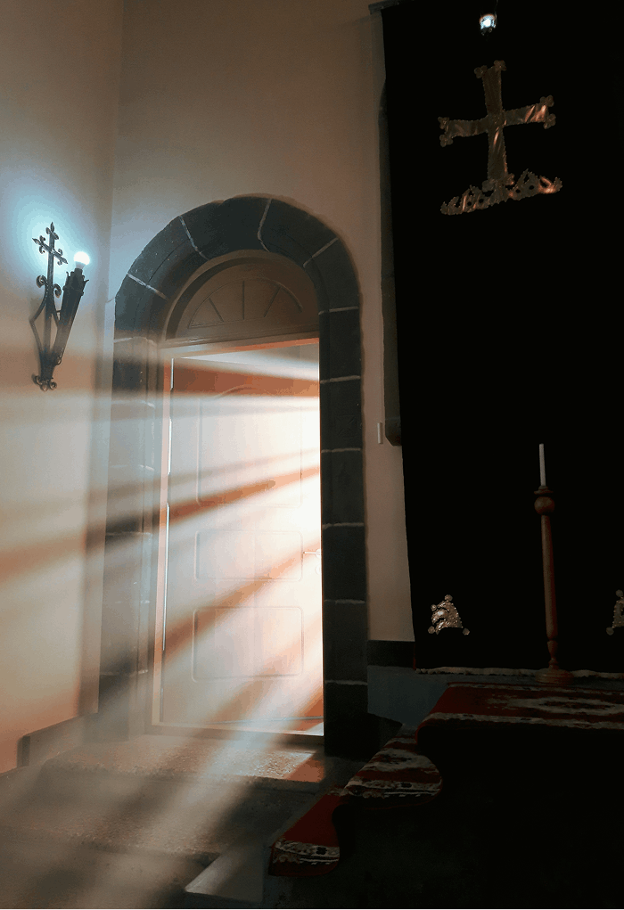 Sunlight streaming through an open door, casting light and shadows inside a room with religious decor, including a cross on the wall and a candle holder.
