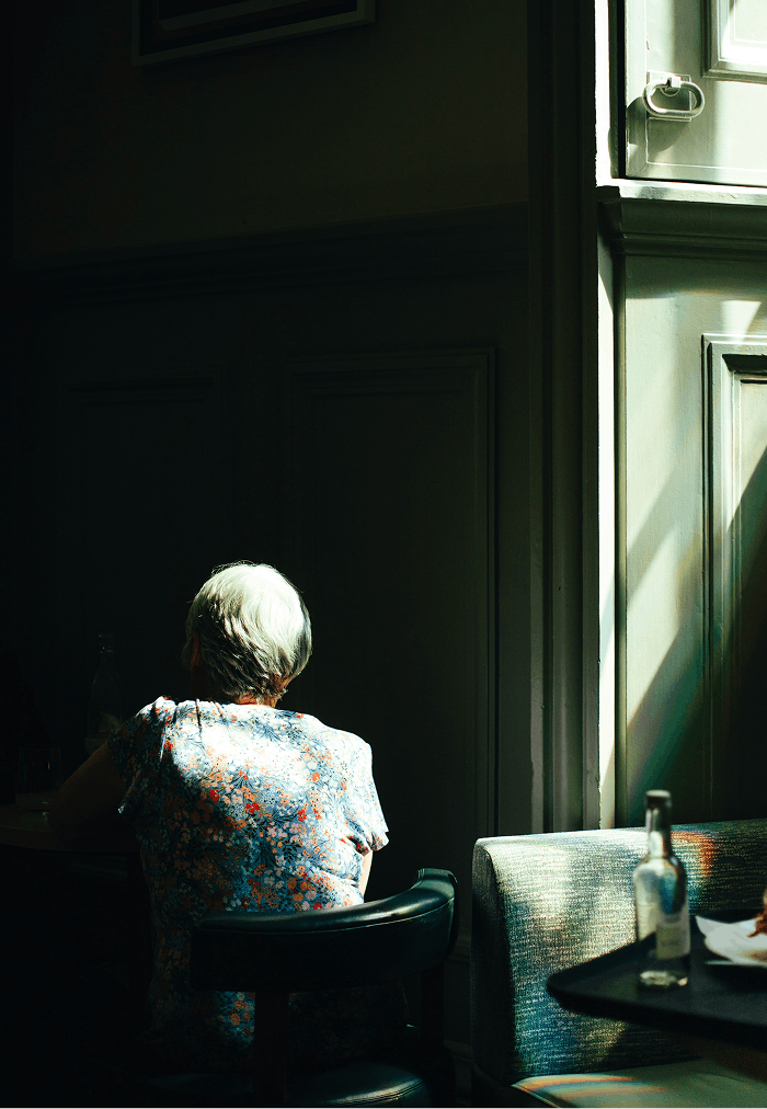 Older woman with short gray hair sitting alone at a table in a dimly lit room, illuminated by sunlight coming through a window.