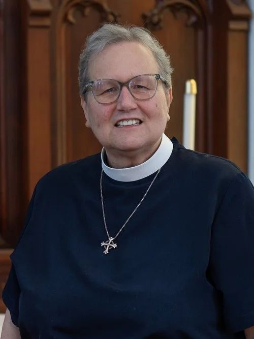A smiling woman dressed in clergy attire, wearing glasses and a cross necklace, standing indoors with wooden paneling and a lit candle in the background.