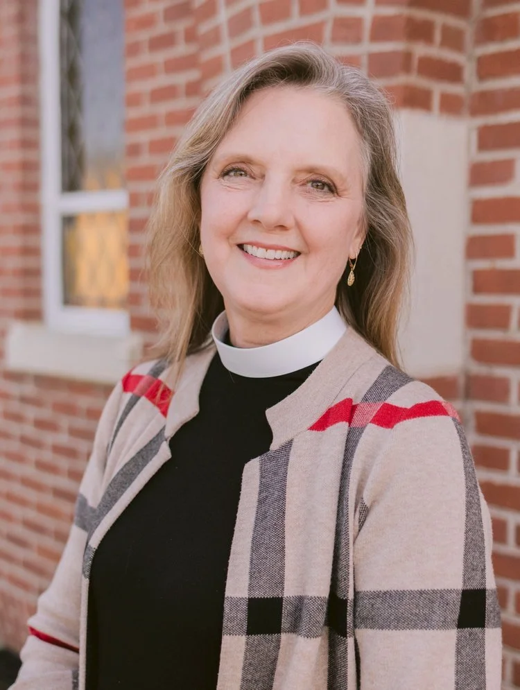 A smiling woman wearing a clergy collar and a plaid jacket standing outside in front of a brick building.