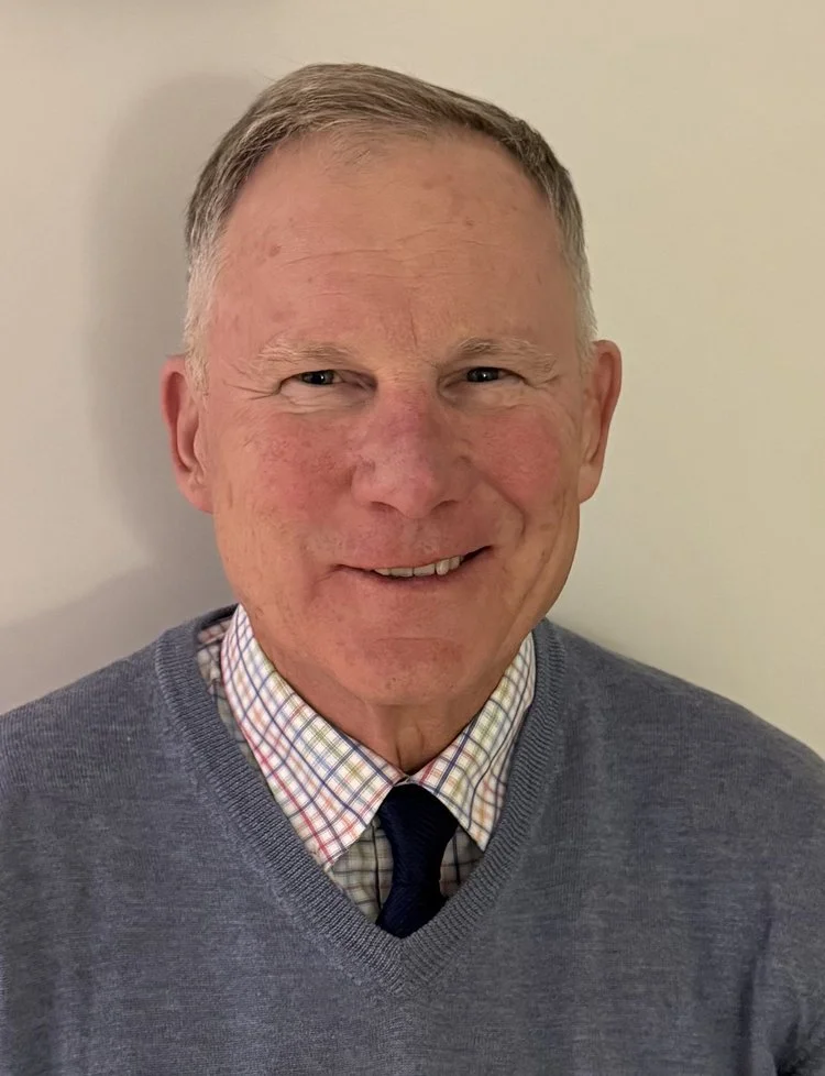 A smiling man with short gray hair, wearing a checkered dress shirt, a tie, and a gray sweater, standing against a plain light-colored wall.