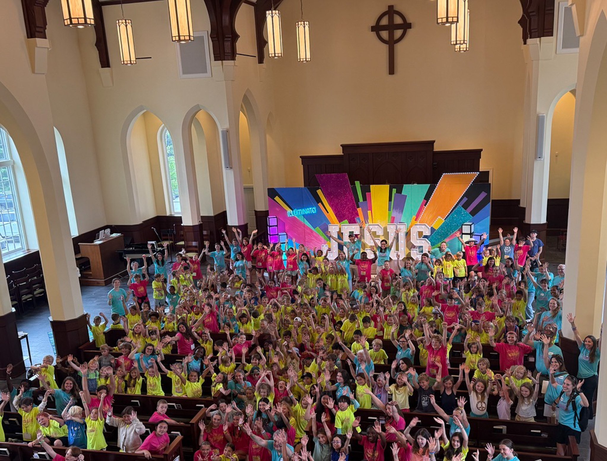 A large group of children and adults gathered inside a church, many wearing bright-colored shirts, waving and smiling at the camera, with a stage decorated with colorful banners and a cross on the wall behind them.