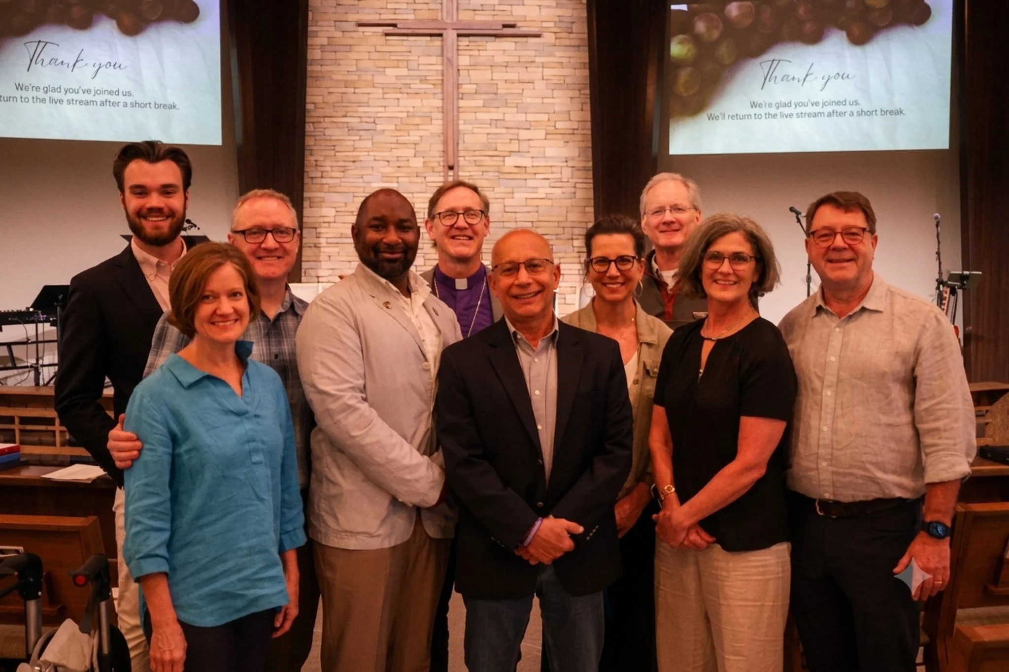 A group of ten people standing in front of a brick wall with a cross, smiling for a photo after a church service. Two screens behind them display a thank you message.