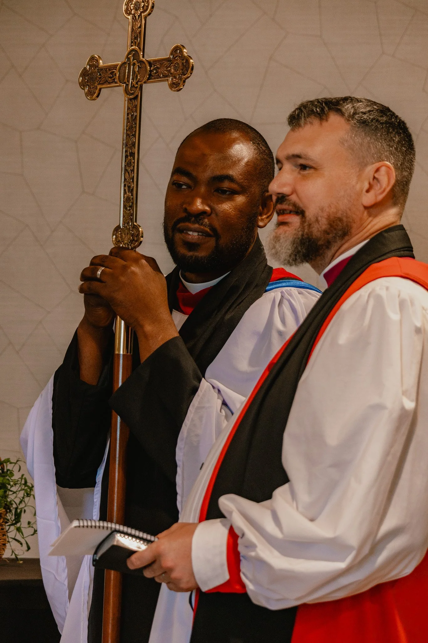 Two men in religious attire, one holding a large ornate cross, standing inside a church. The man on the left is black, and the man on the right has light skin and a beard.