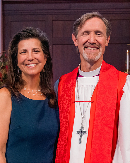 A woman in a blue dress and a priest in clerical attire with a red stole posing together indoors.