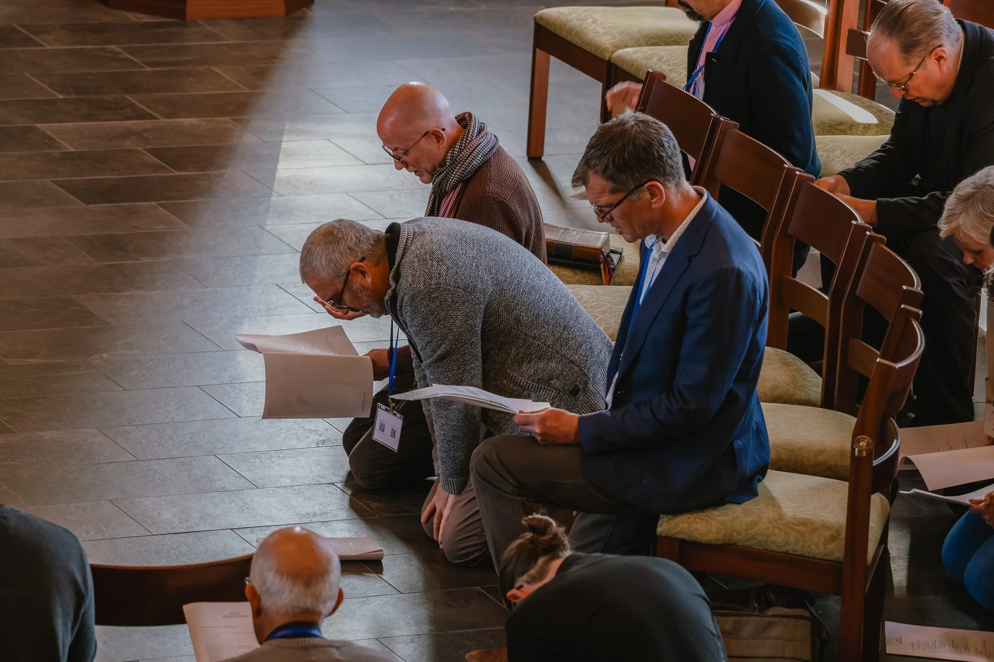 People kneeling and praying during a religious service or gathering in a church or hall, with some holding sheets of paper or hymnals.