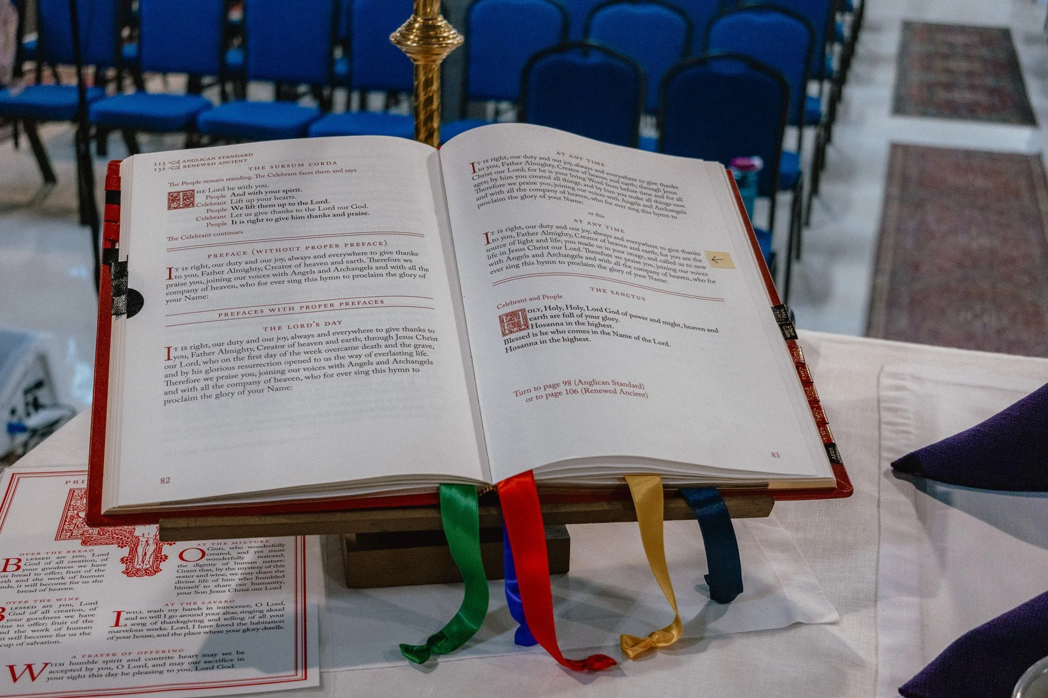 Open Bible resting on a stand with multiple colorful ribbons hanging from it. There are rows of blue chairs in the background, indicating a church or religious setting.