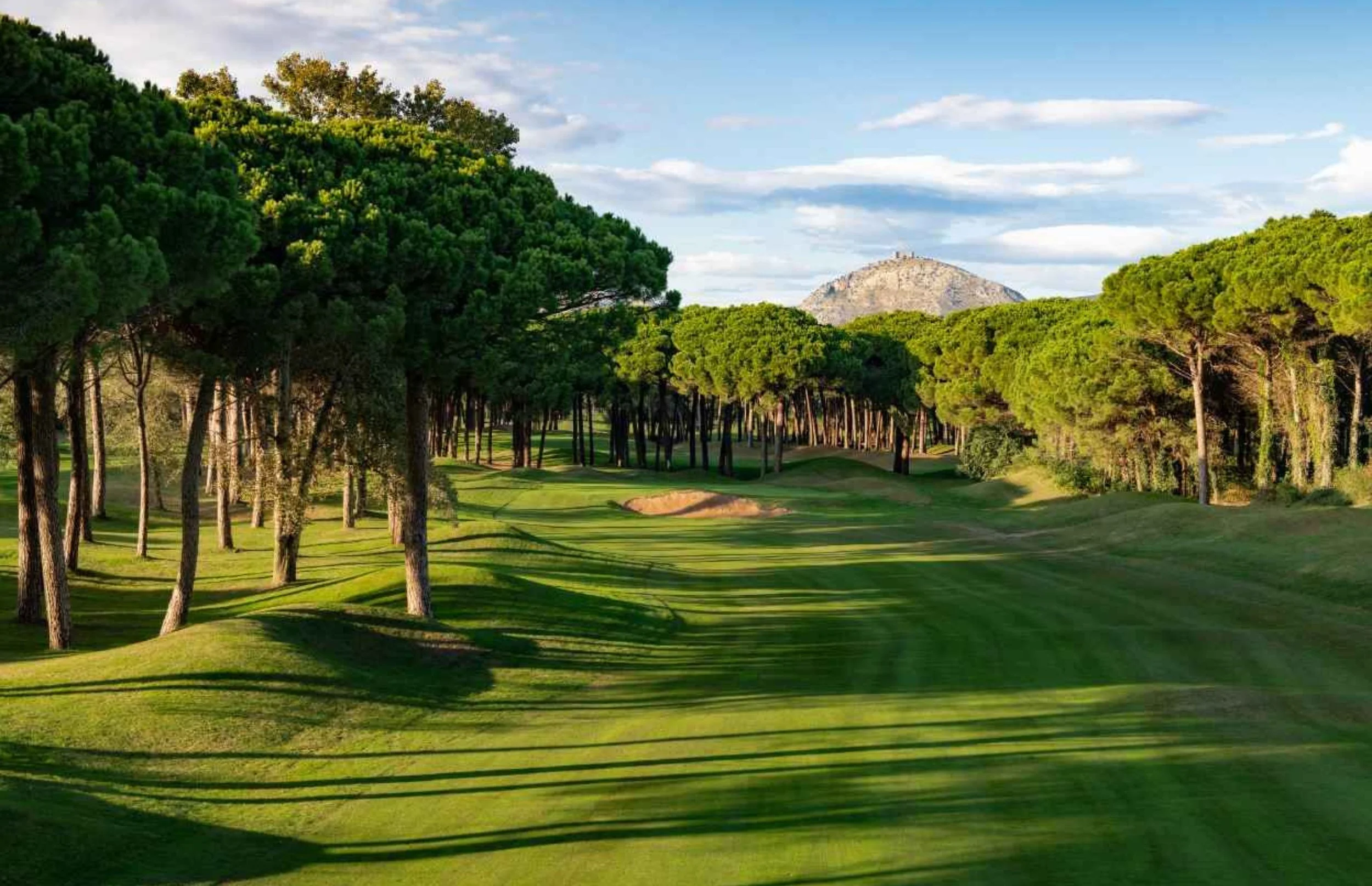 A golf course with green fairways and tall trees lining the sides, with a mountain in the background and a partly cloudy sky above.