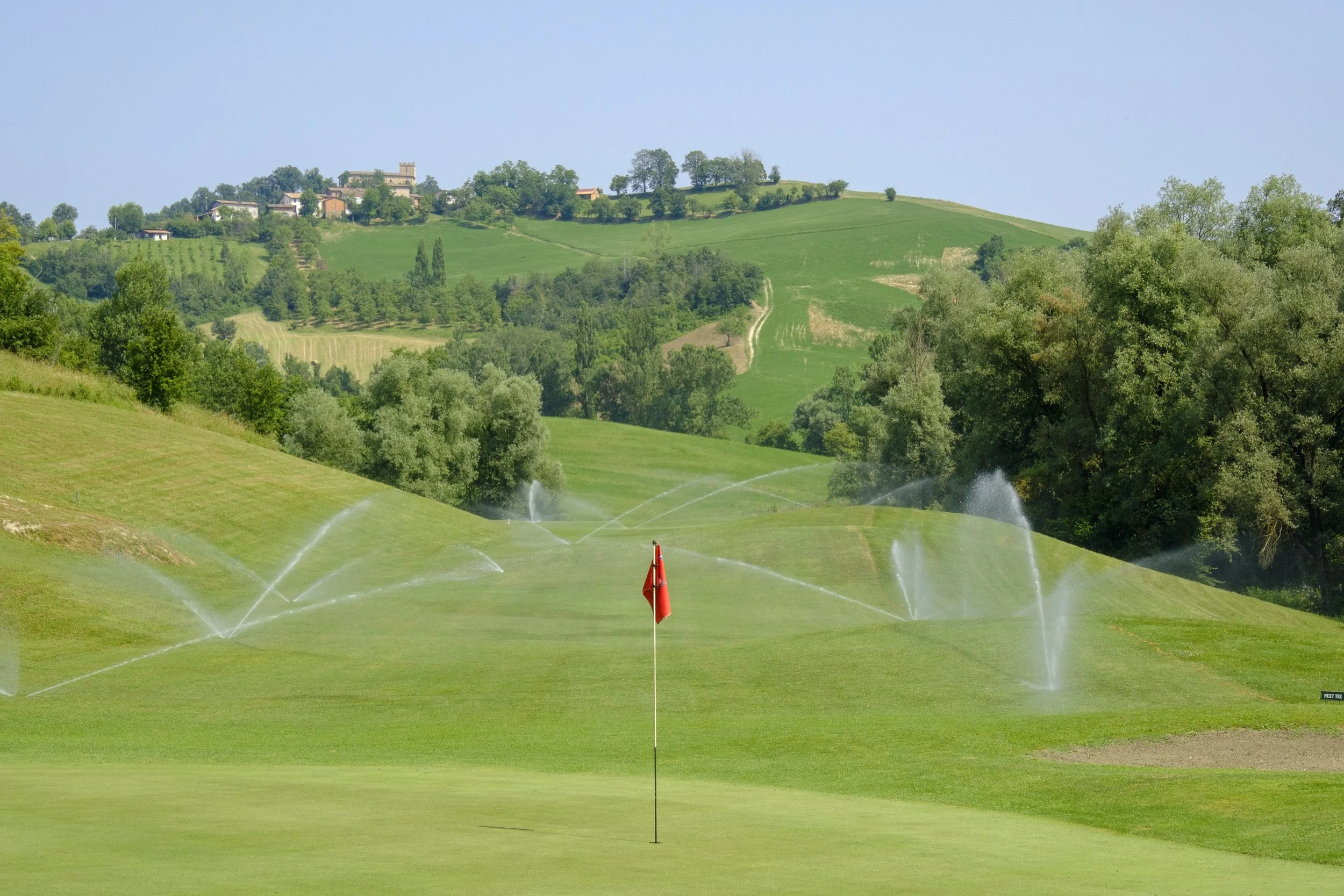 A golf course with sprinklers watering the green, a red flag on the putting green, and rolling hills with houses and trees in the background under a clear blue sky.