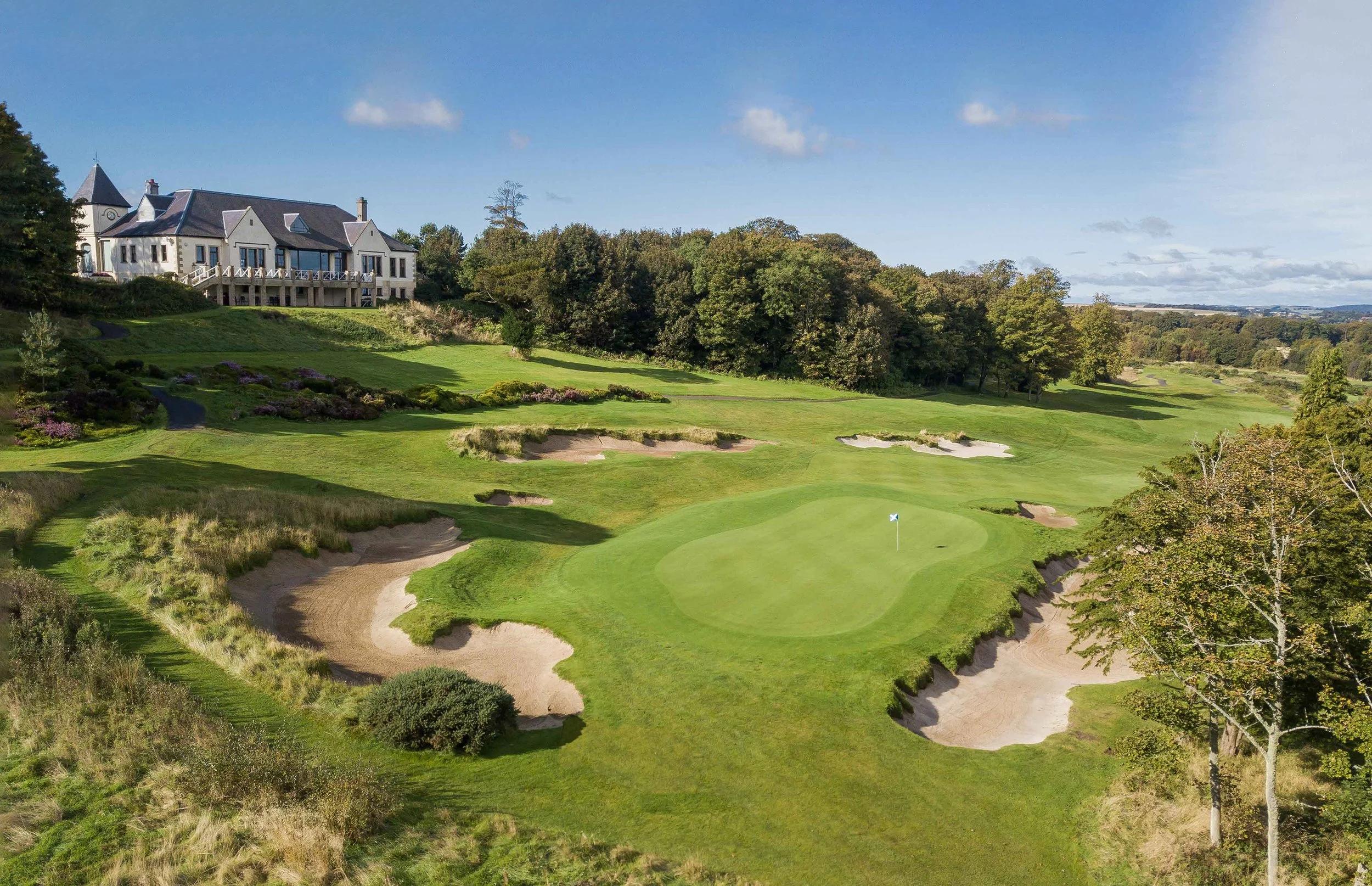 Scenic golf course with green, sand traps, trees, and a large house on a hill in the background under a blue sky.