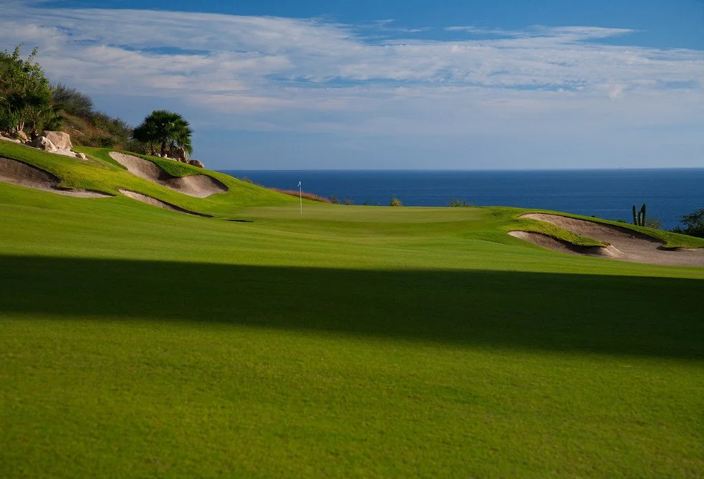 A golf course with green grass, sand bunkers, a flagstick, and ocean in the background under a partly cloudy sky.