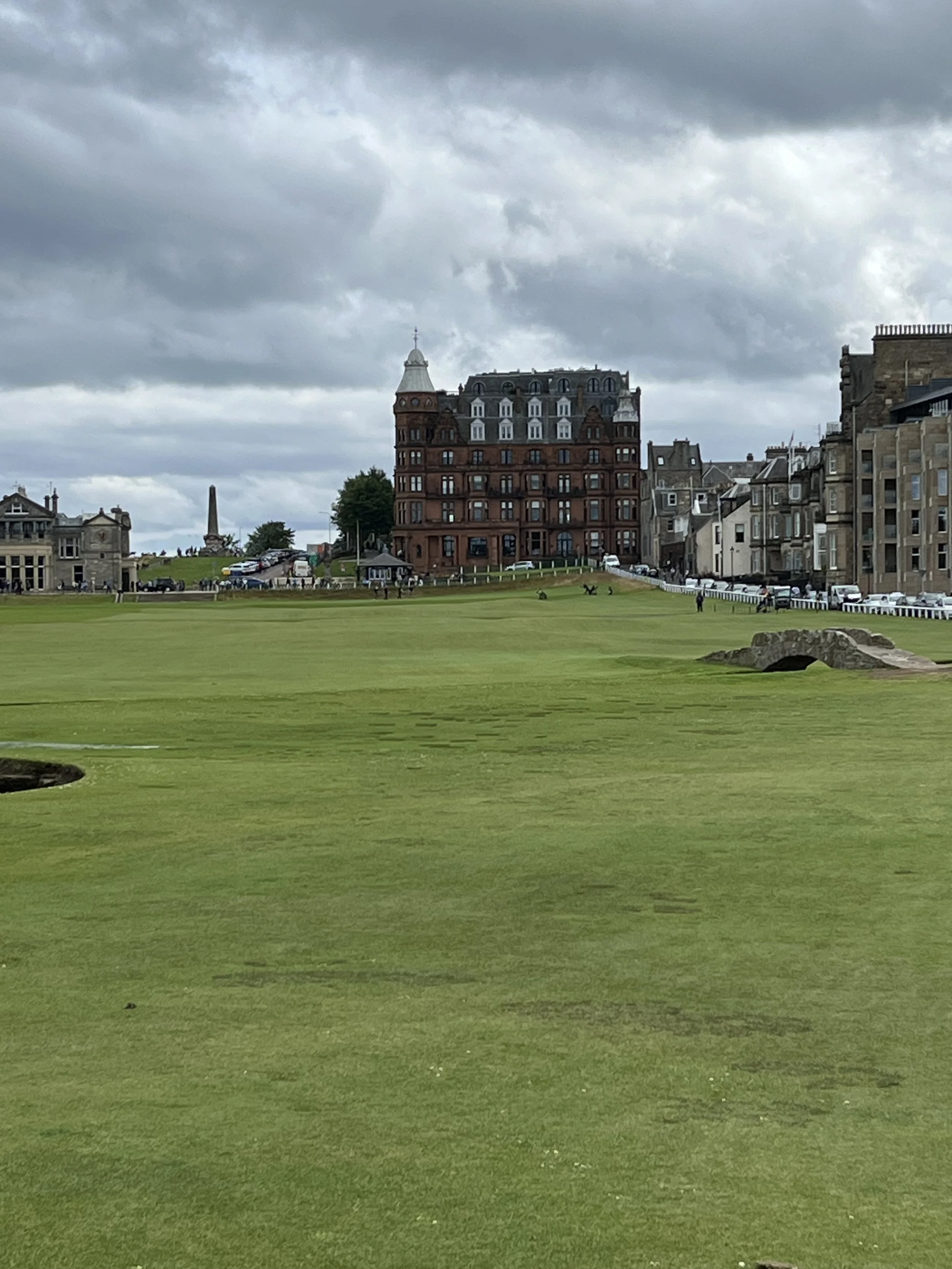 A golf course with green grass under a cloudy sky, with large historic buildings and a monument in the background.