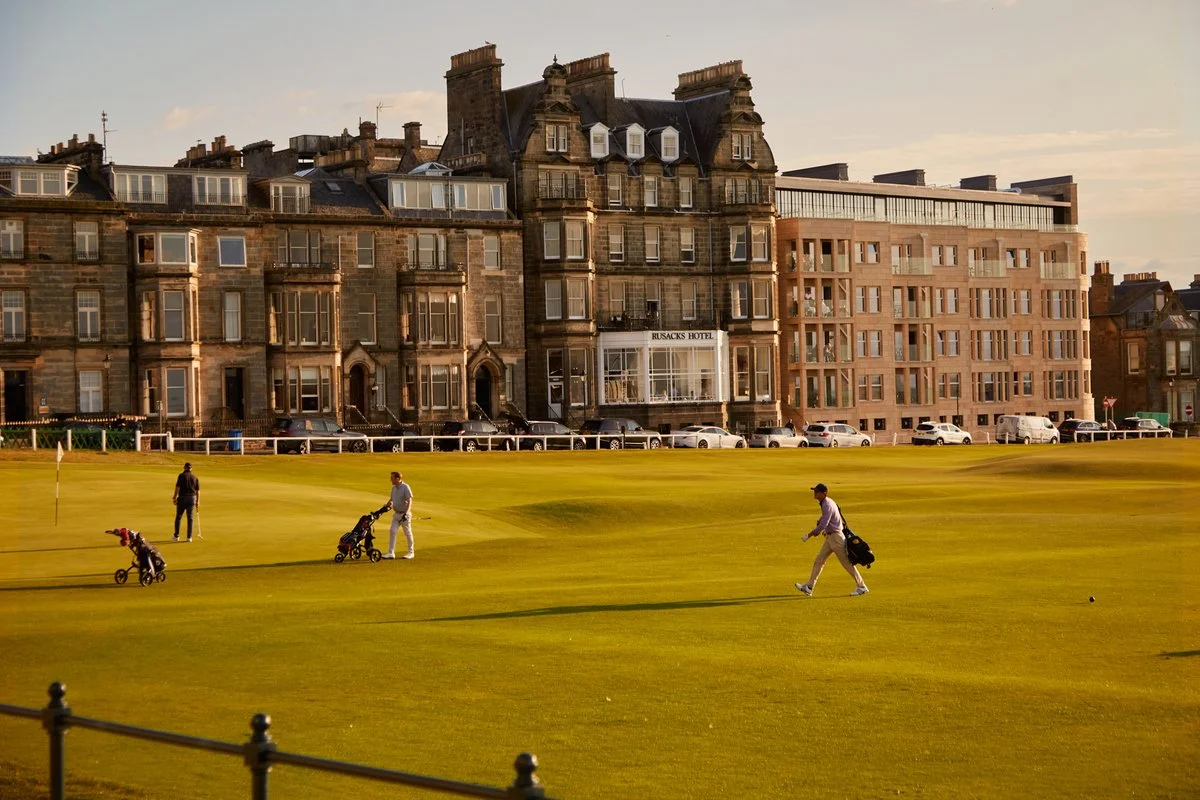 Golfers walking on a golf course with bags and carts, with historic stone buildings in the background during sunset.