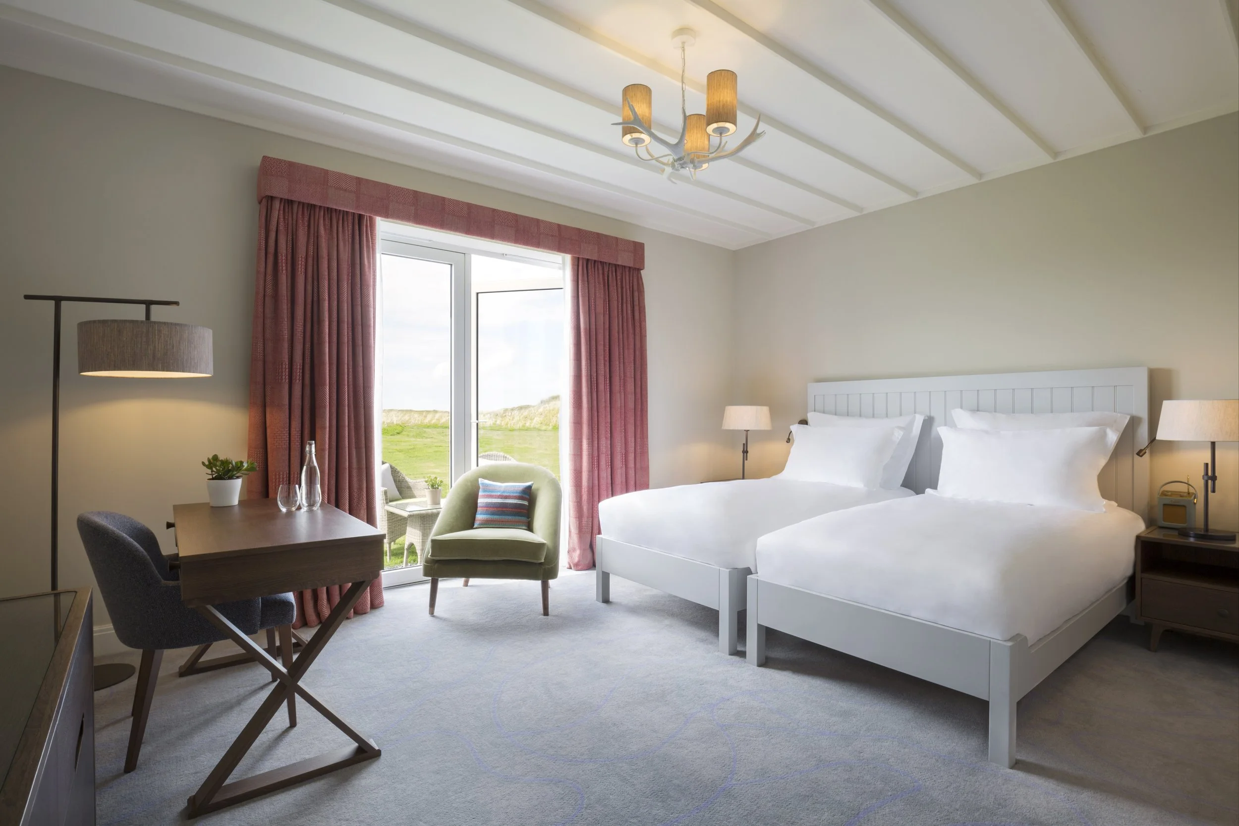 Hotel room with two beds, a sitting area near a window, pink curtains, a desk with a chair, and a view of grassy landscape outside.