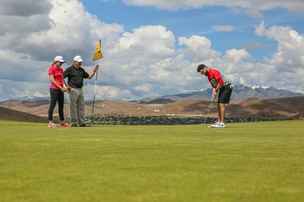Two women and a man on a golf course, with one man preparing to putt, and an instructor holding a flag