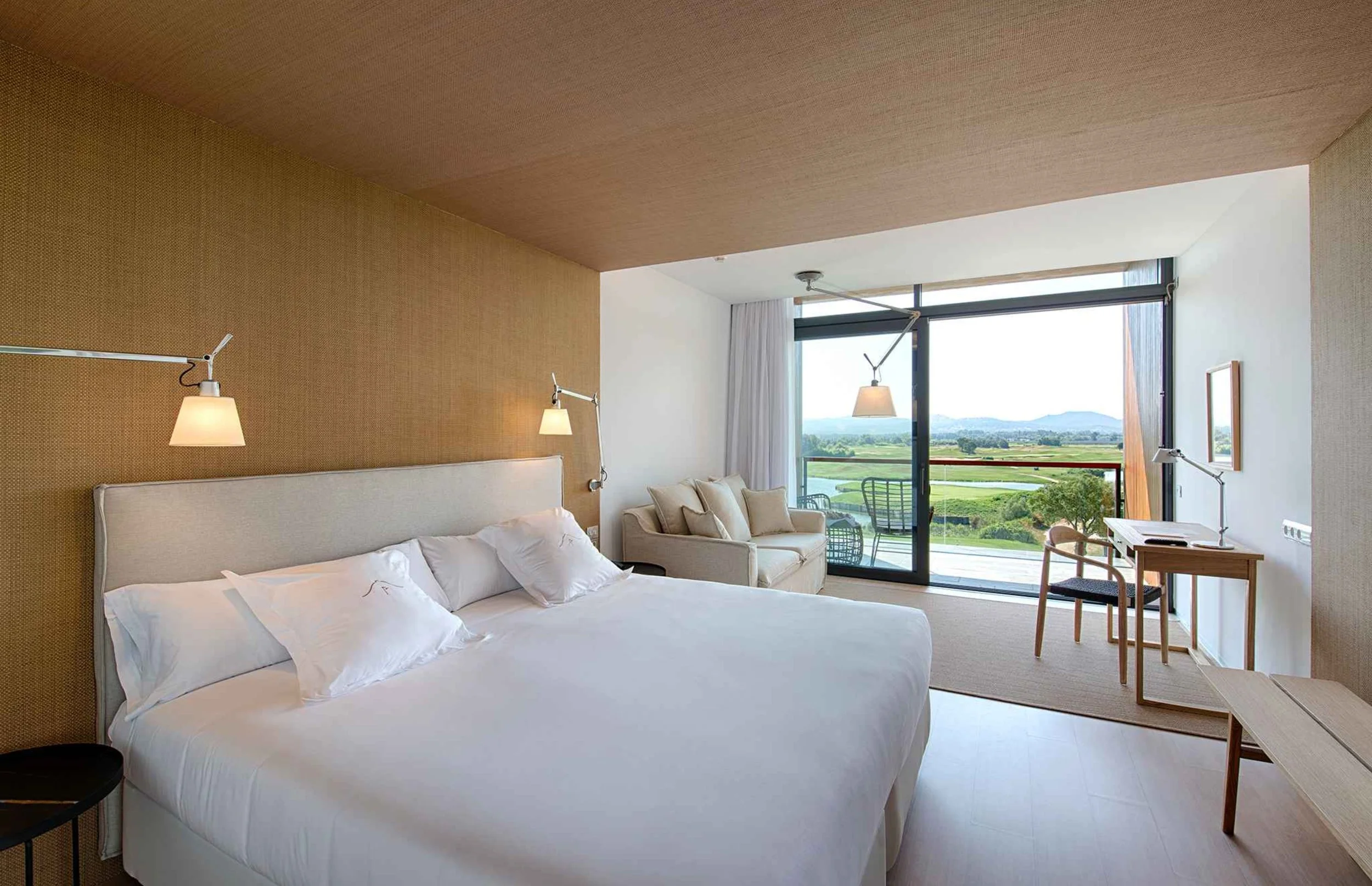 A modern hotel room with a large bed, white bedding, beige headboard, and wall lamps. There is a sitting area with a sofa and pillows, a wooden desk with a chair, and a sliding glass door leading to a balcony with a scenic view of green fields, water