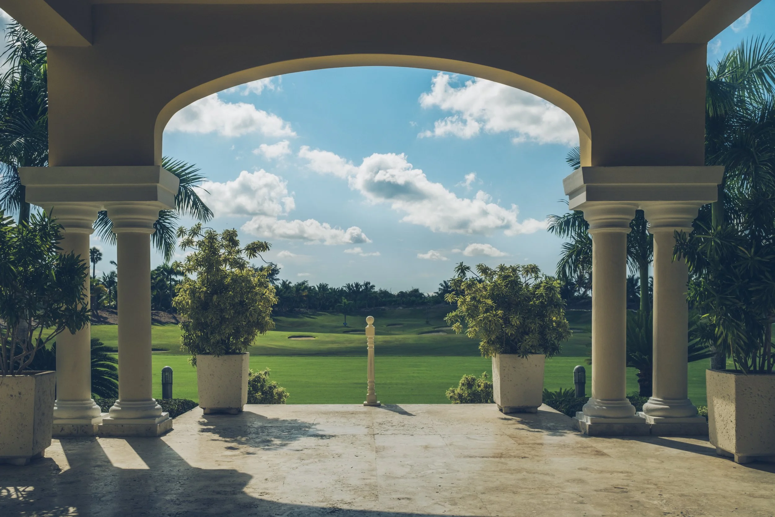View from a veranda with white columns, overlooking a lush golf course in a tropical setting with palm trees, under a partly cloudy blue sky.