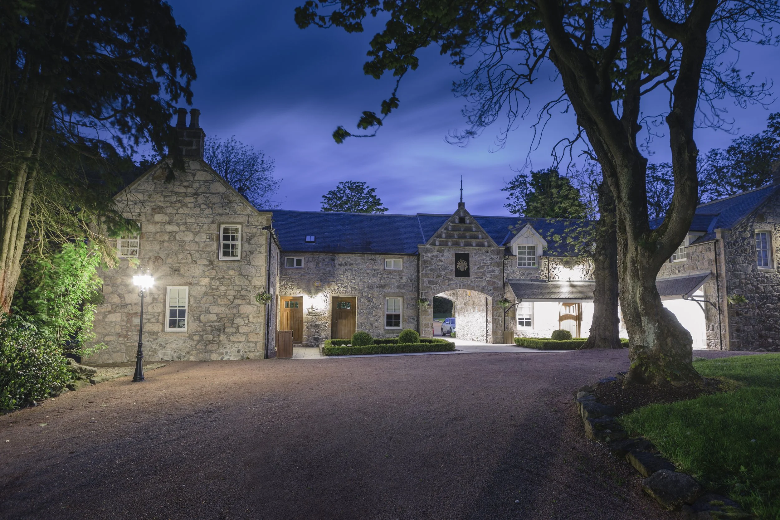 A stone building with arched entryways and multiple windows, surrounded by trees and a gravel driveway, illuminated at dusk.
