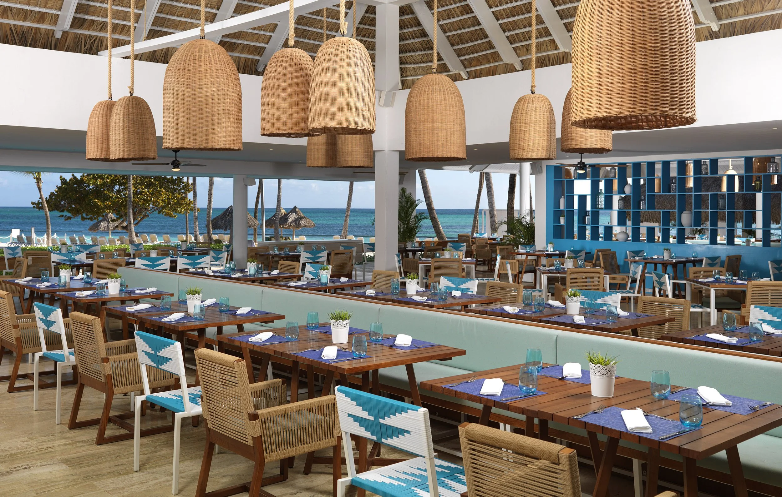Beachside restaurant with wooden tables and chairs, blue glassware, and potted plants, overlooking the ocean with palm trees and thatched umbrellas in the background.