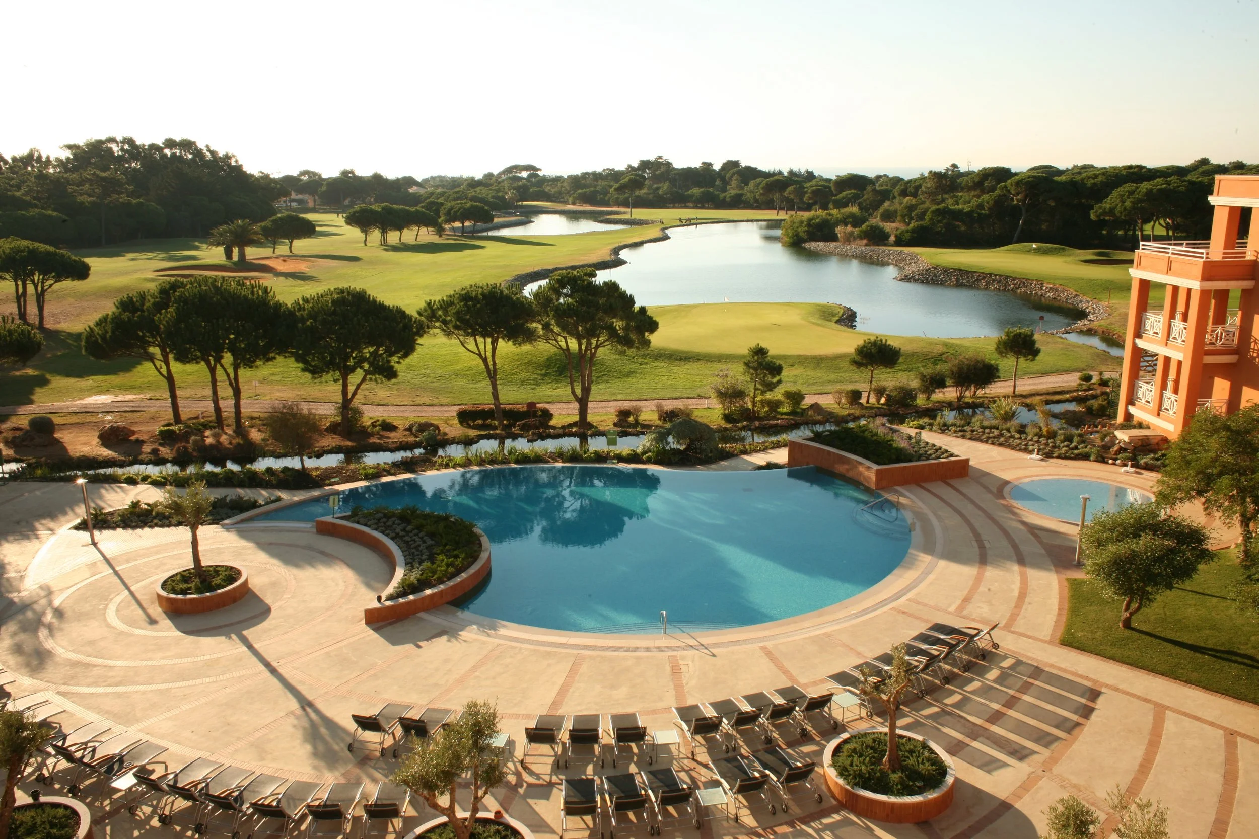 View of a swimming pool with lounge chairs, overlooking a scenic golf course with water features, trees, and a lush landscape, under a clear sky.