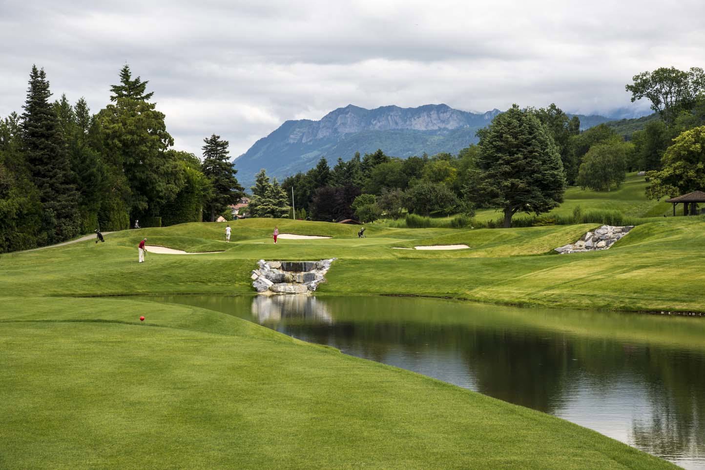 Golfers play on a lush green course featuring a stone waterfall and pond, set against the breathtaking mountain peaks of the Rhône-Alpes region during a luxury golf vacation in France.