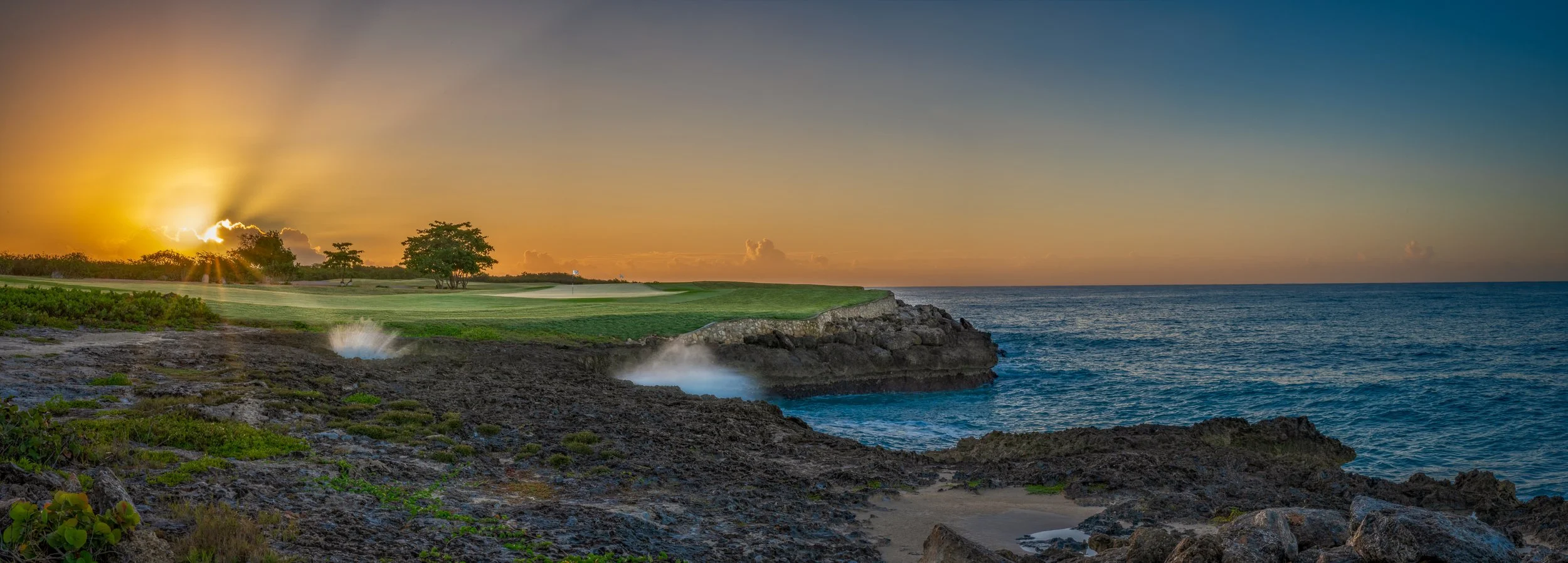 Sunset over a coastal golf course with a putting green, trees, and rocky shoreline by the ocean.