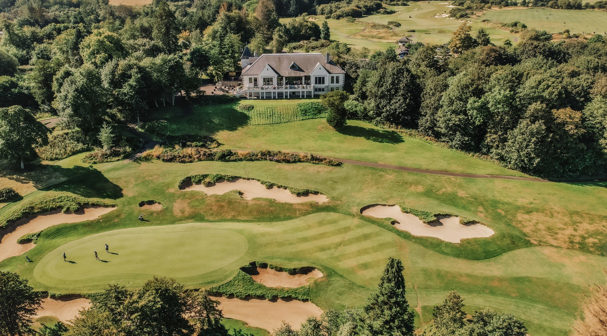 Aerial view of a golf course with sand traps, a putting green, and a clubhouse surrounded by trees and fields.