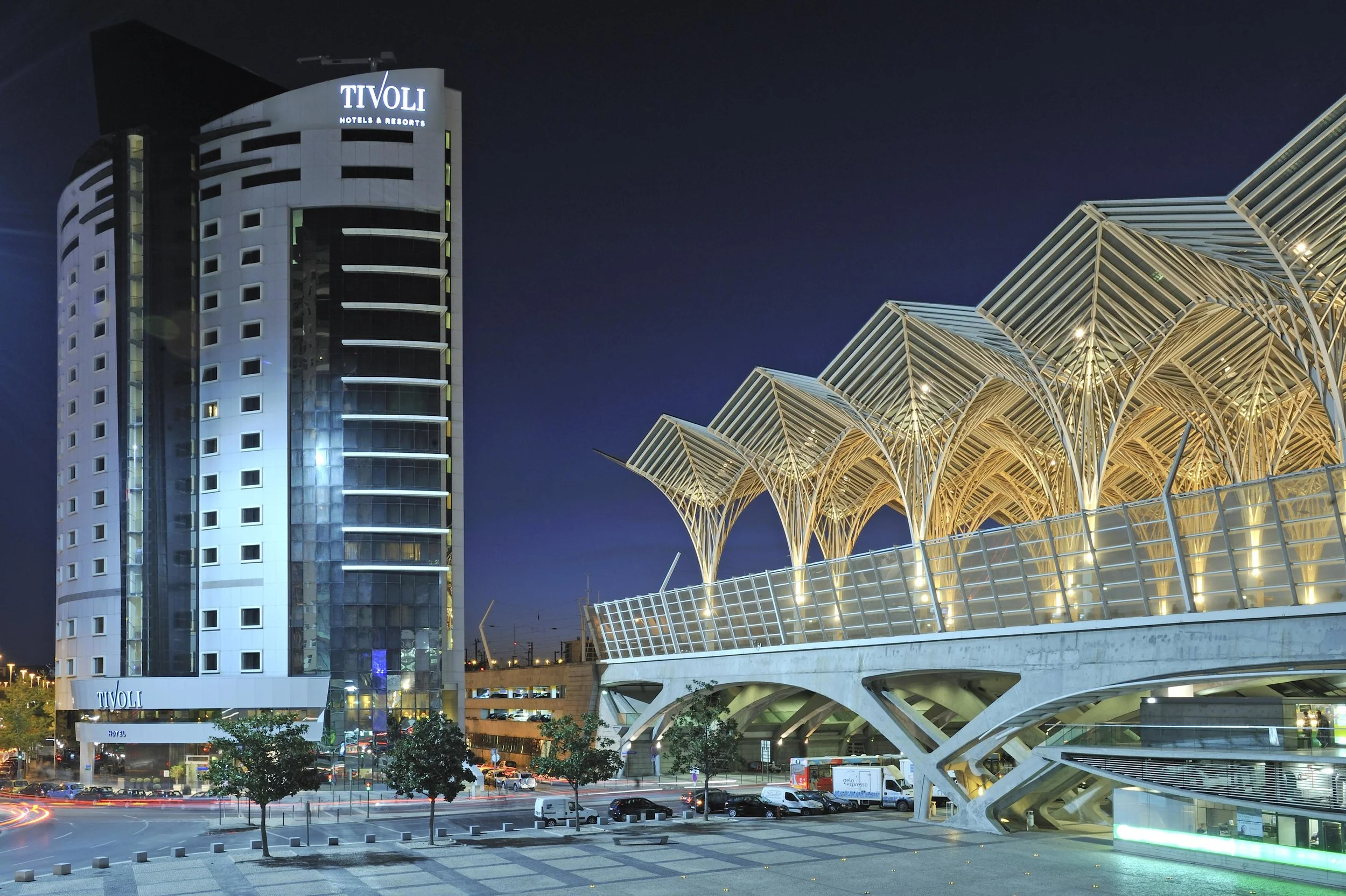 Night view of a modern cityscape featuring a tall building with the sign Tivoli Hotels & Resorts and a unique architectural structure with ribbed, curved roofs, illuminated against a dark sky.