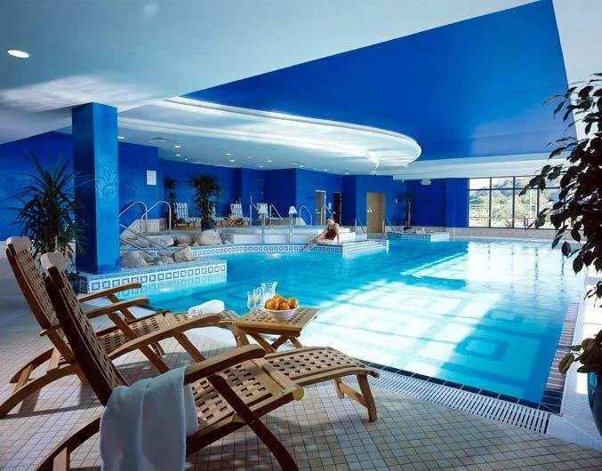 Indoor swimming pool area with wooden lounge chairs, a tray of drinks, and a person relaxing in the pool, with blue walls and large windows letting in natural light.