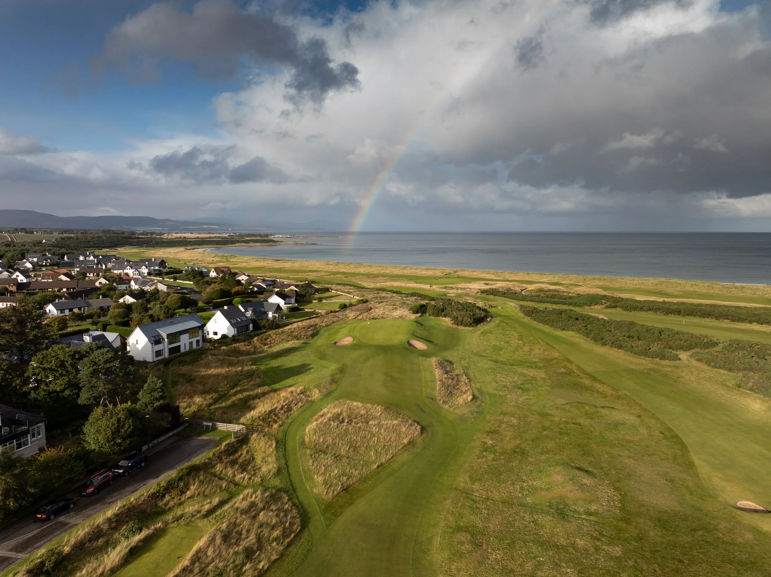Aerial view of a coastal golf course with a rainbow in the sky over the ocean and a neighborhood of white houses nearby.