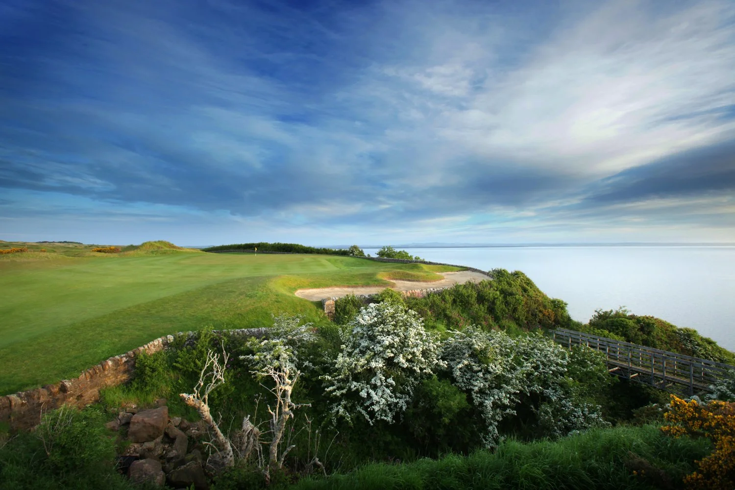 A scenic view of a golf course situated on a hillside overlooking a body of water, with lush green grass, flowering bushes, and a wooden footbridge against a cloudy sky.