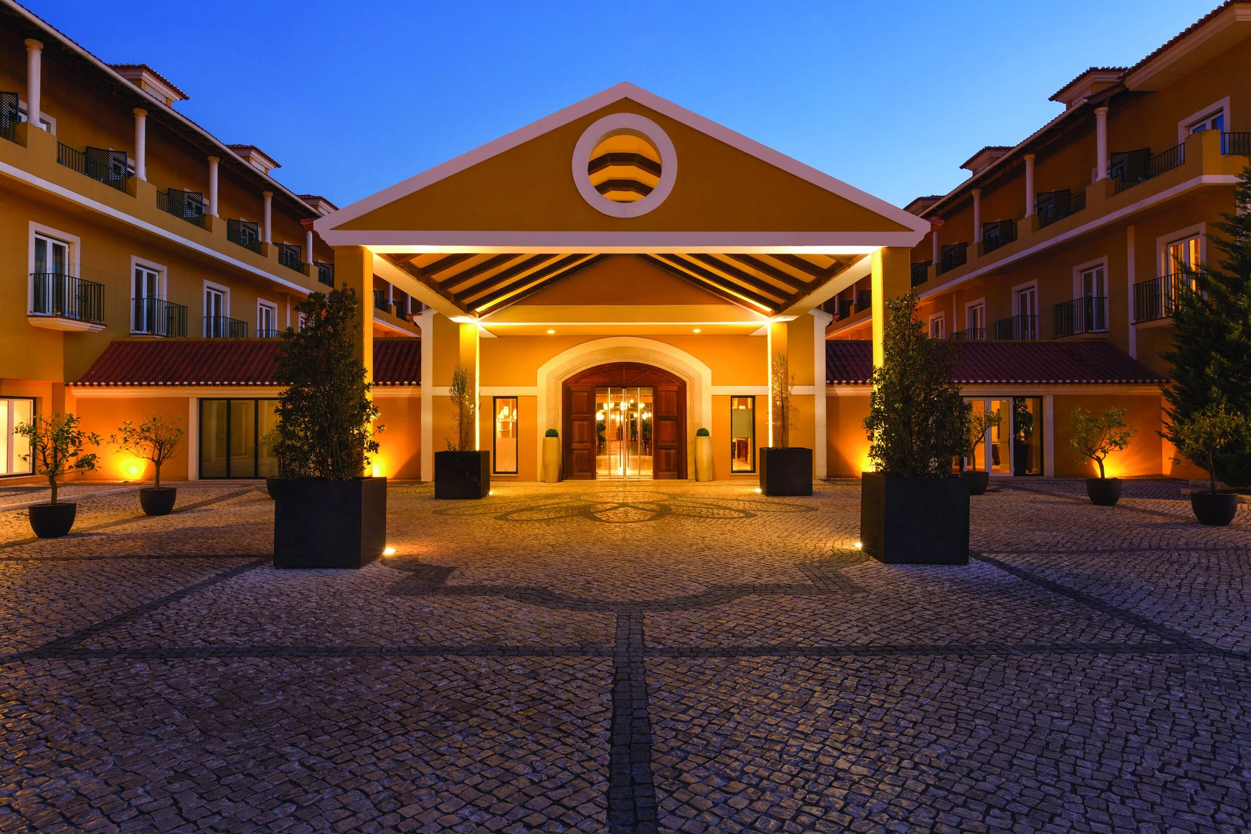 Entrance of a well-lit hotel or resort during dusk, with a cobblestone driveway, potted plants, and a modern building with balconies on each side.