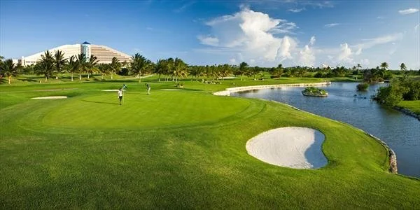 A golf course with players, a water hazard, and a city building in the background under a partly cloudy sky.