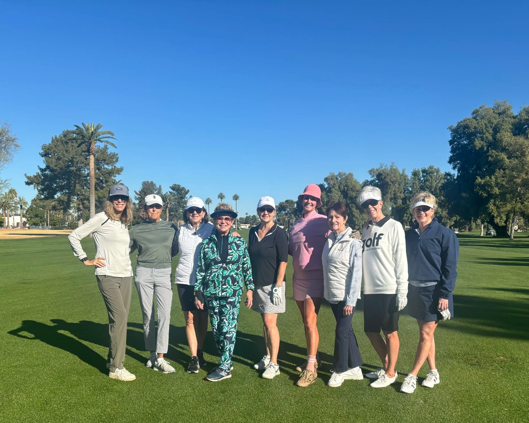 Group of nine women standing together on a golf course under a clear blue sky, dressed in golf attire and smiling.
