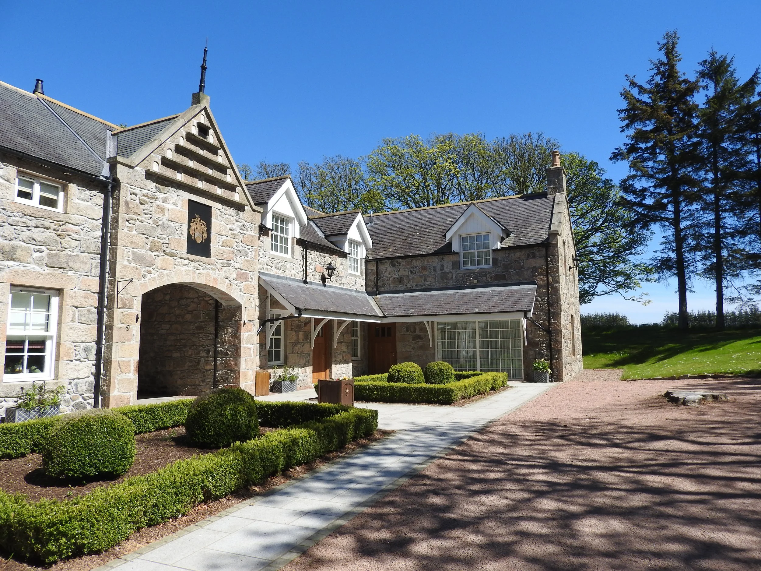 A stone house with dormer windows and a covered entrance, surrounded by trimmed bushes and a gravel yard, under a clear blue sky.