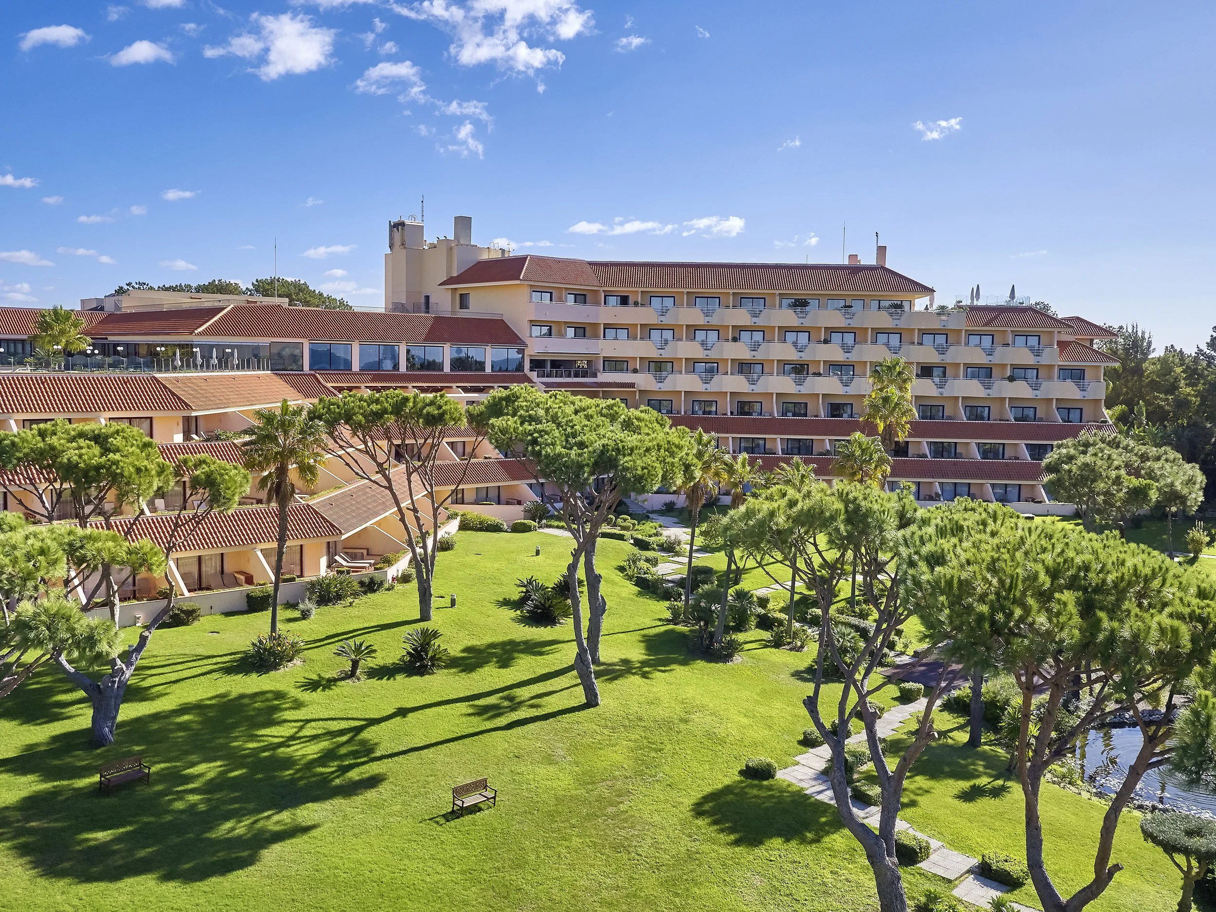 A large hotel or resort building with multiple floors and balconies, surrounded by green trees, well-manicured grass, and a pathway, under a blue sky with some clouds.