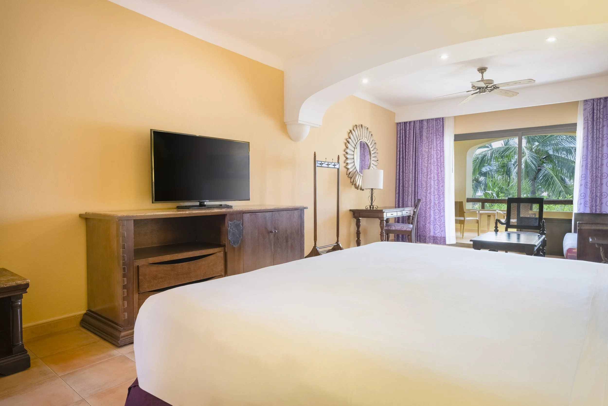Hotel room with a bed, a wooden TV stand with a flat-screen TV, a small desk with a lamp and mirror, purple curtains, and a sitting area with a chair and balcony view of palm trees.
