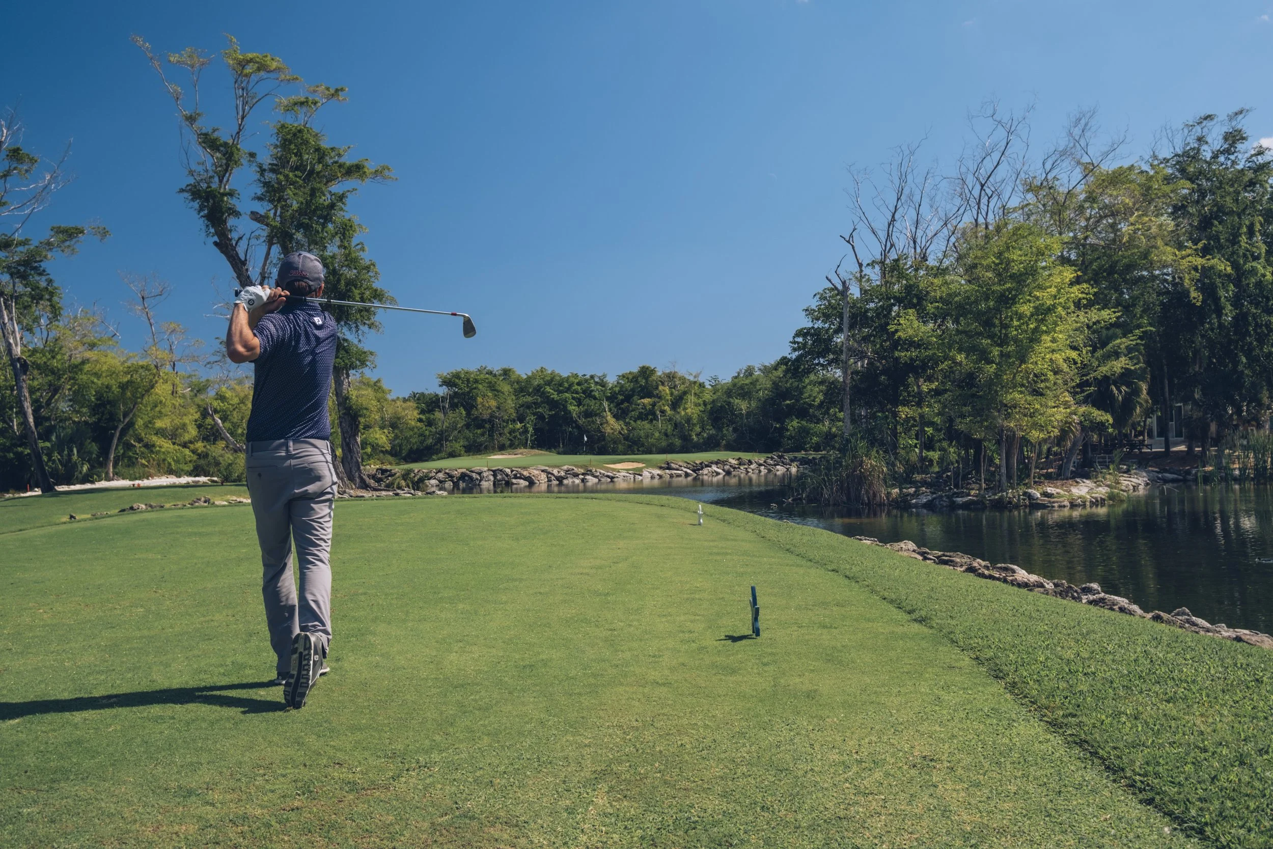 A man playing golf on a lush green course beside a water body with trees in the background under a clear blue sky.