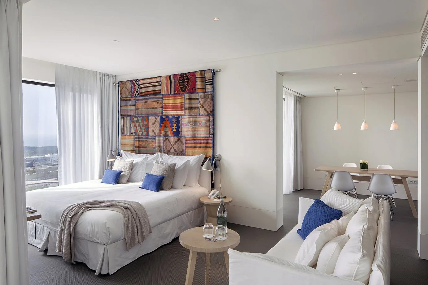 Hotel room with a large bed, colorful textile wall hanging, white and blue pillows, a small round table with water bottles and glasses, a sofa with white and blue pillows, and a dining area with three pendant lights and white chairs.