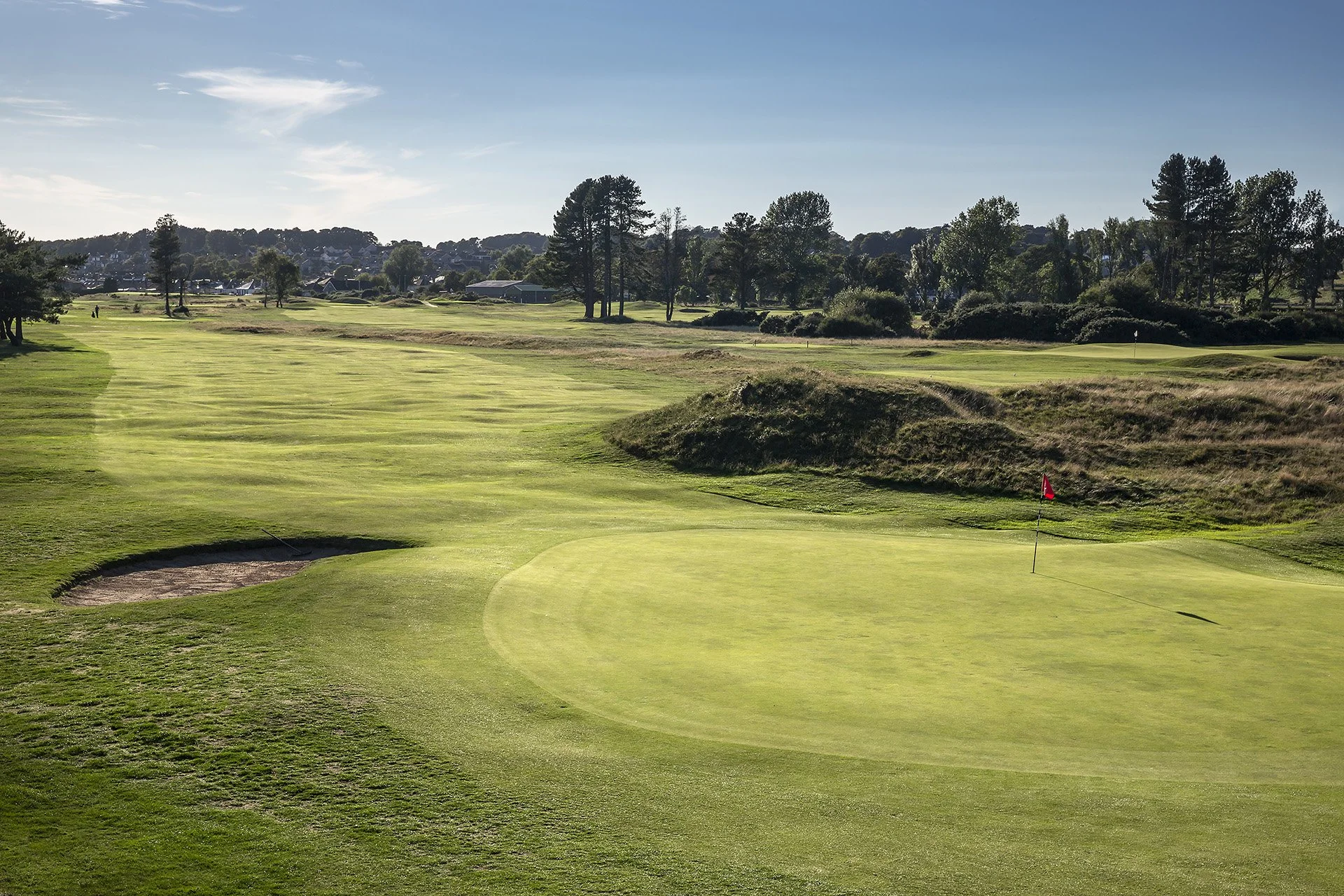 A scenic golf course with green fairways, sand traps, trees, and distant houses under a clear blue sky.