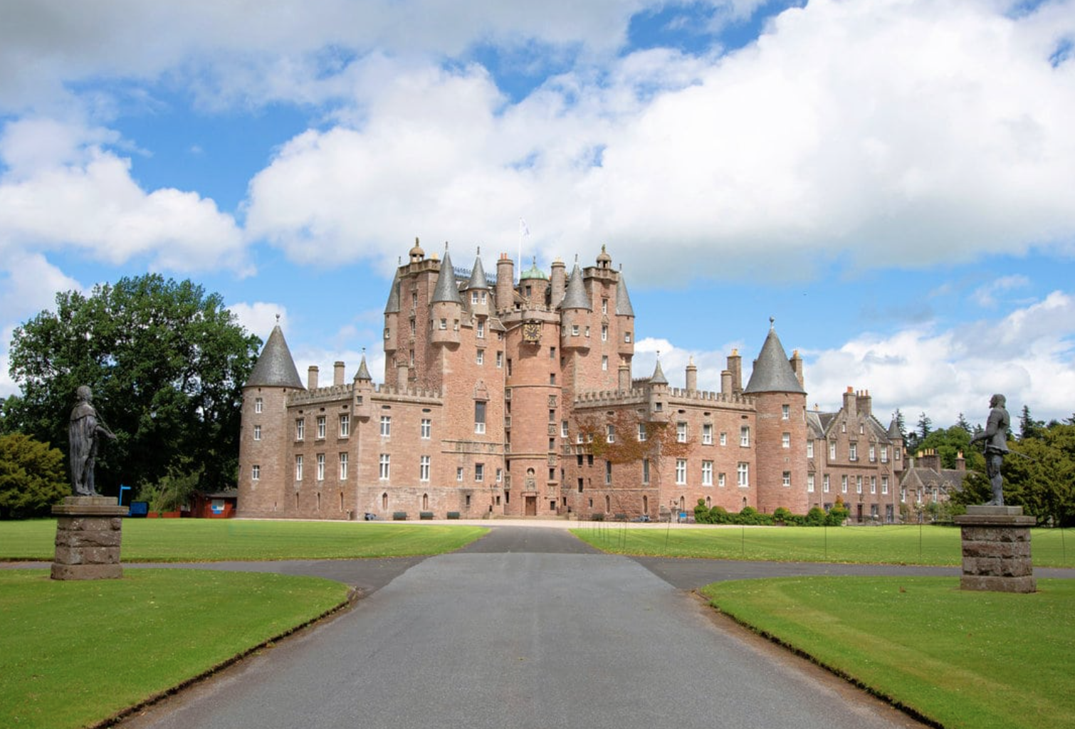 A large stone castle with multiple towers and turrets, surrounded by green lawns and statues, under a partly cloudy sky.