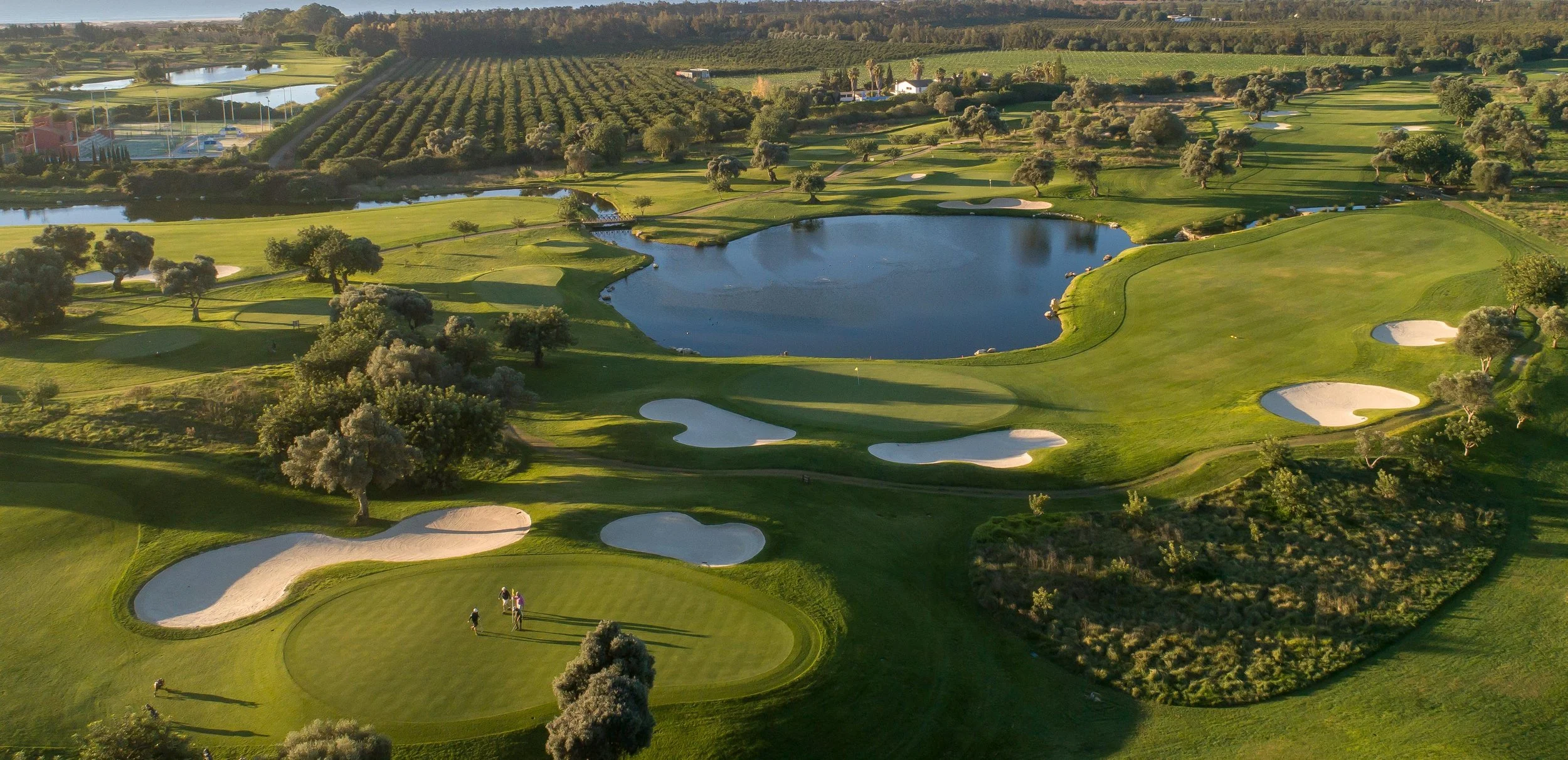 Aerial view of a golf course with several sand traps, water hazards, and a few golfers playing near the green. Surrounding farmland, trees, and small buildings are visible in the background.