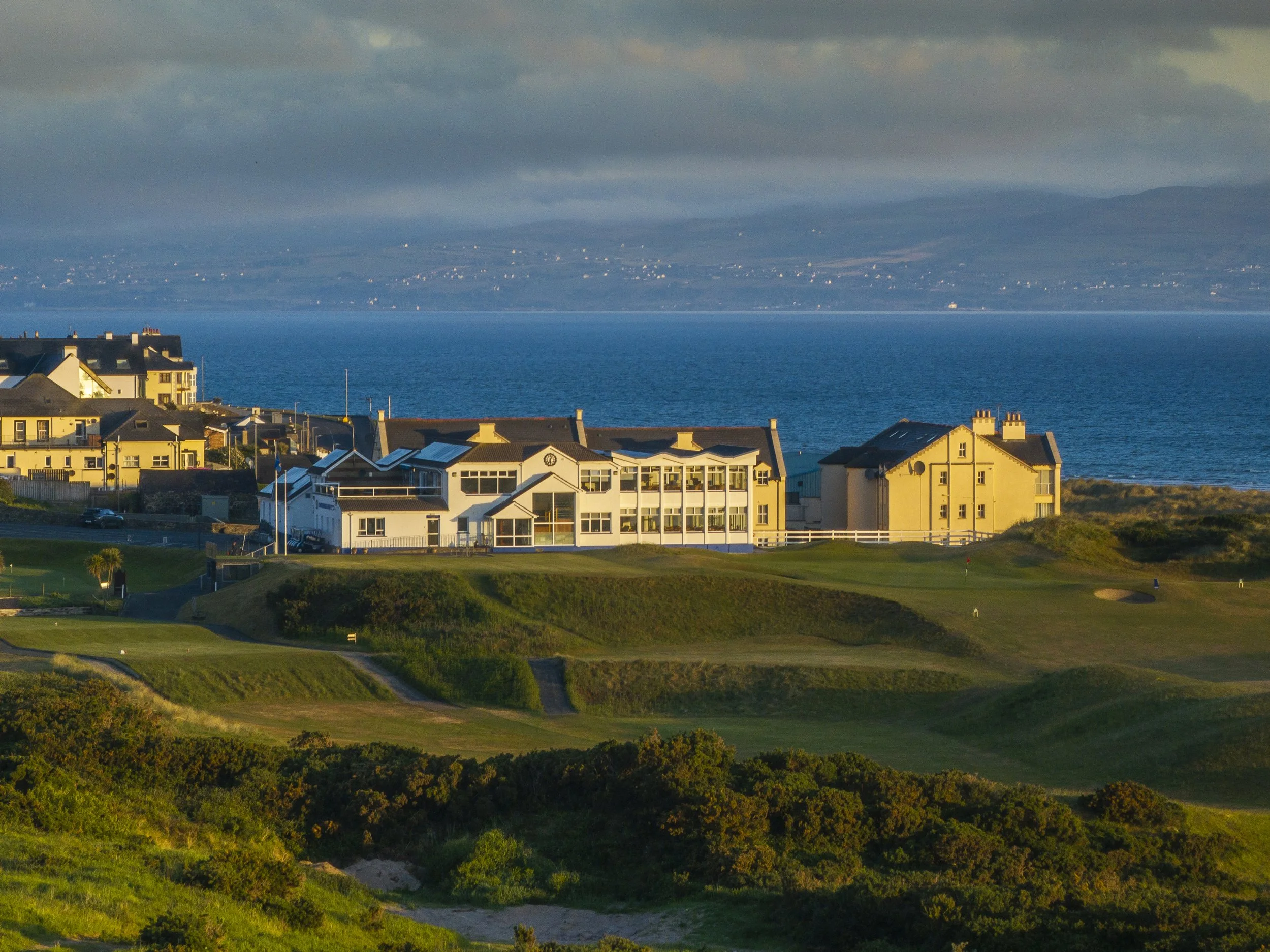 Golf course with green and sand traps, buildings overlooking the ocean, and hills in the background.