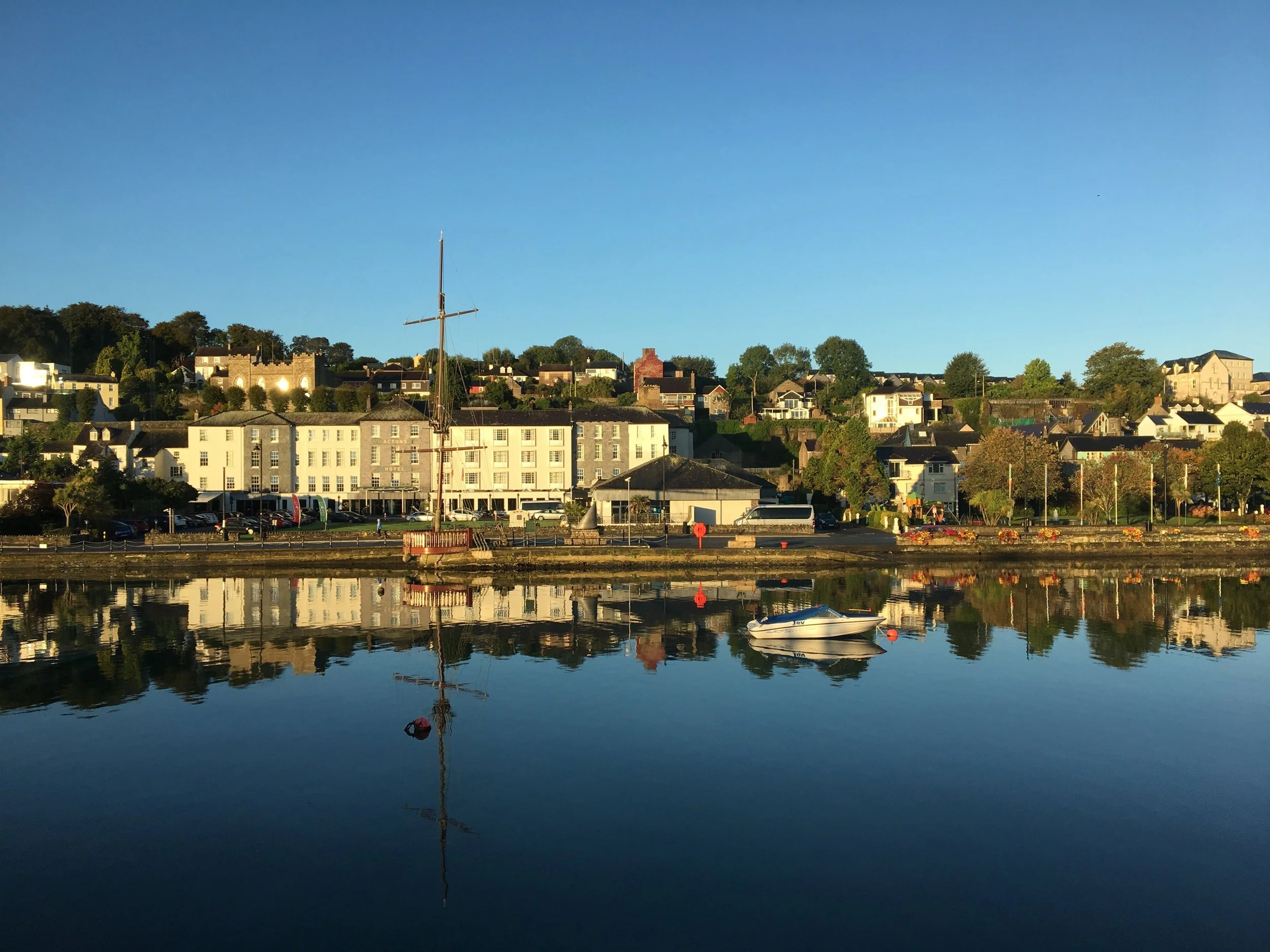 A peaceful waterfront scene with a historic ship docked in the calm water, reflecting the buildings and trees on the shore with a clear blue sky overhead.