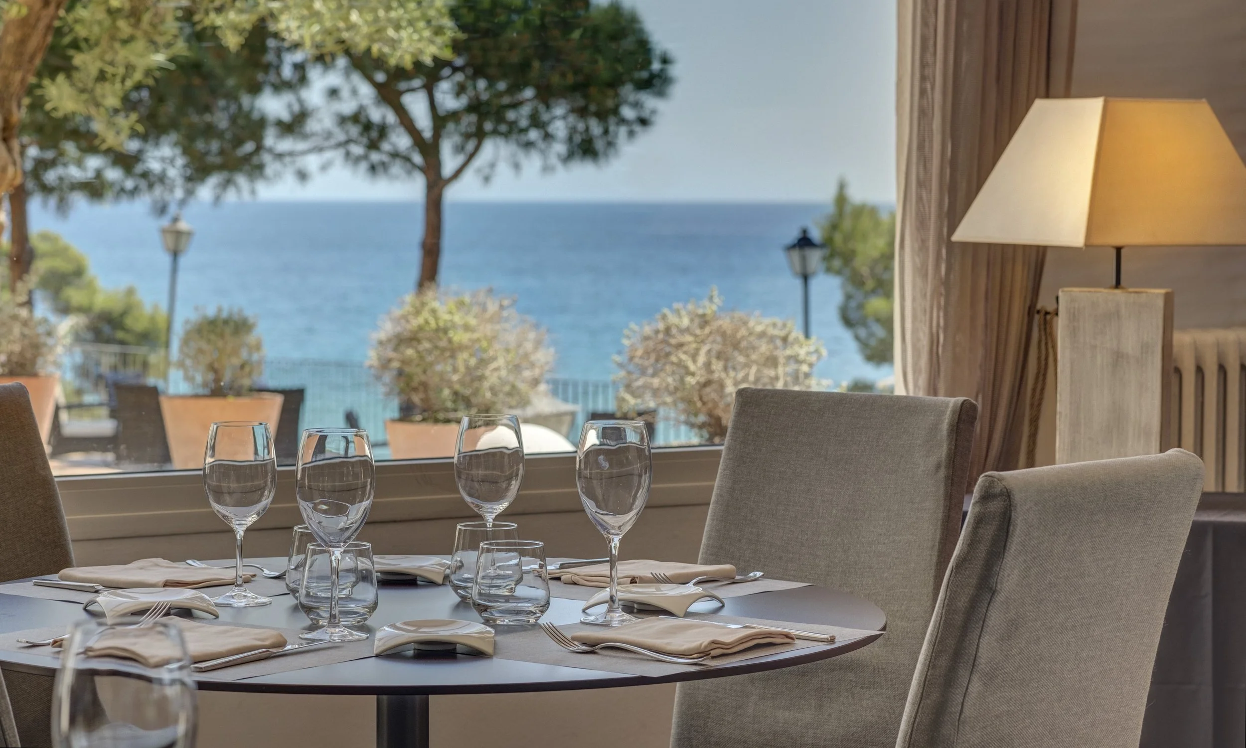 Set dinner table with wine glasses, water glasses, napkins, and silverware indoors, overlooking a view of the ocean and trees outside.
