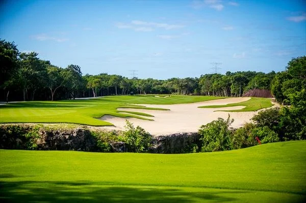 A golf course with lush green grass, sand traps, and trees under a bright blue sky.